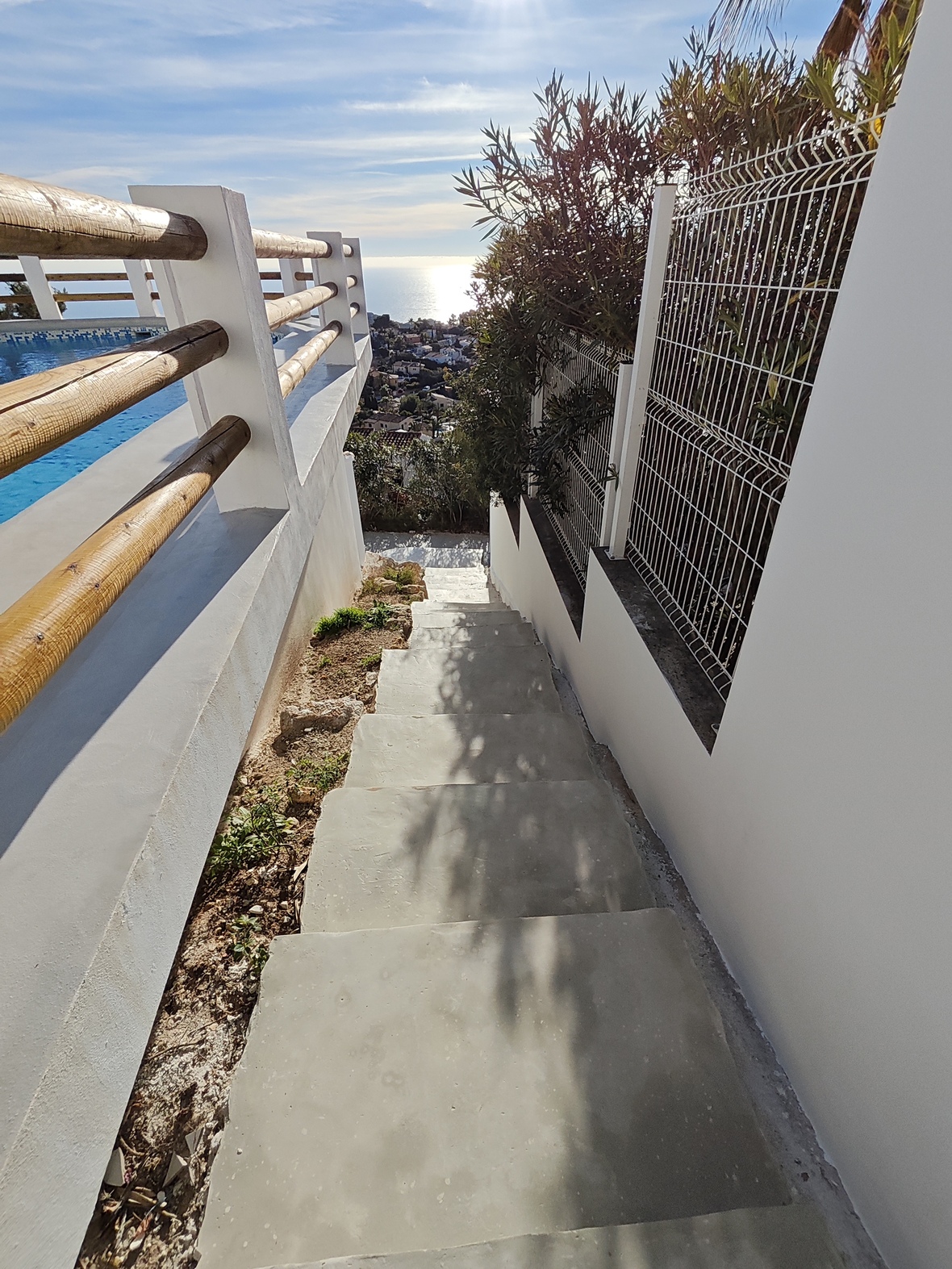 Außentreppe aus Beton mit Holzgeländer und weißen Wänden, die einen Blick auf den Pool und die Mittelmeerküste bei Tageslicht bietet.