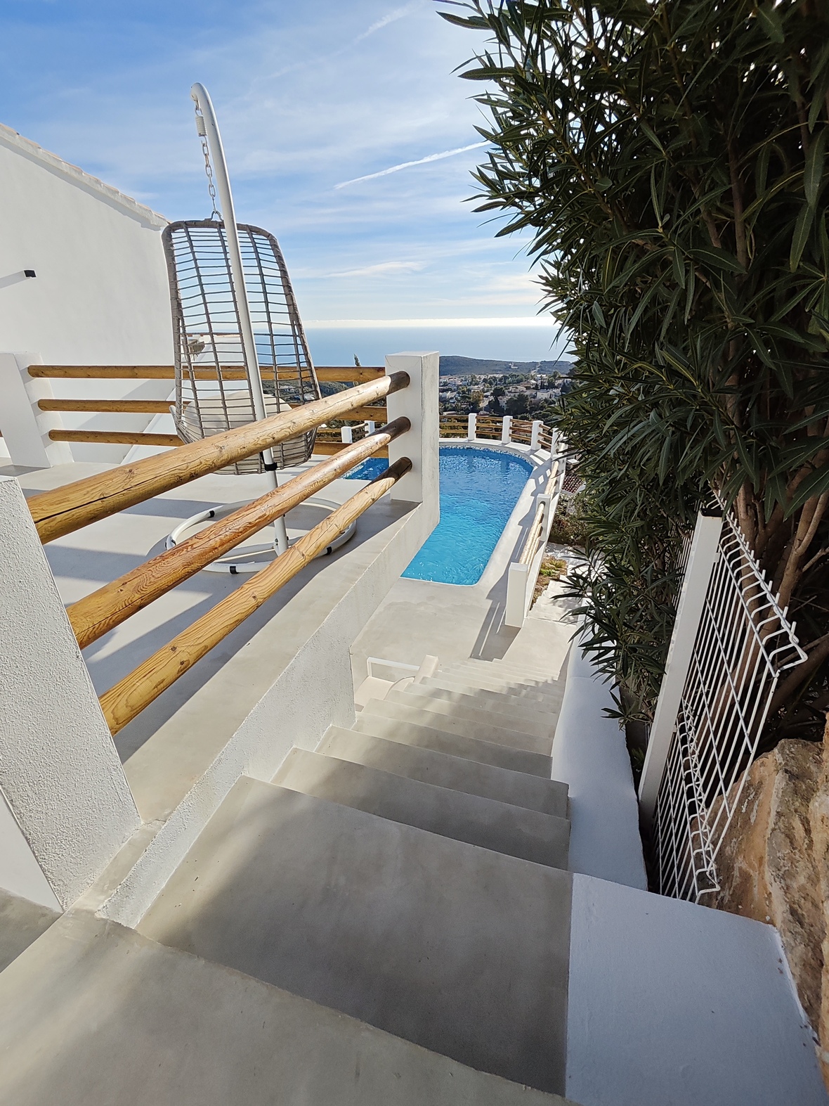 Betontreppen führen zu einer Terrasse mit Hängesessel und Holzgeländer, mit Blick auf den Pool und das Panorama der Mittelmeerküste.