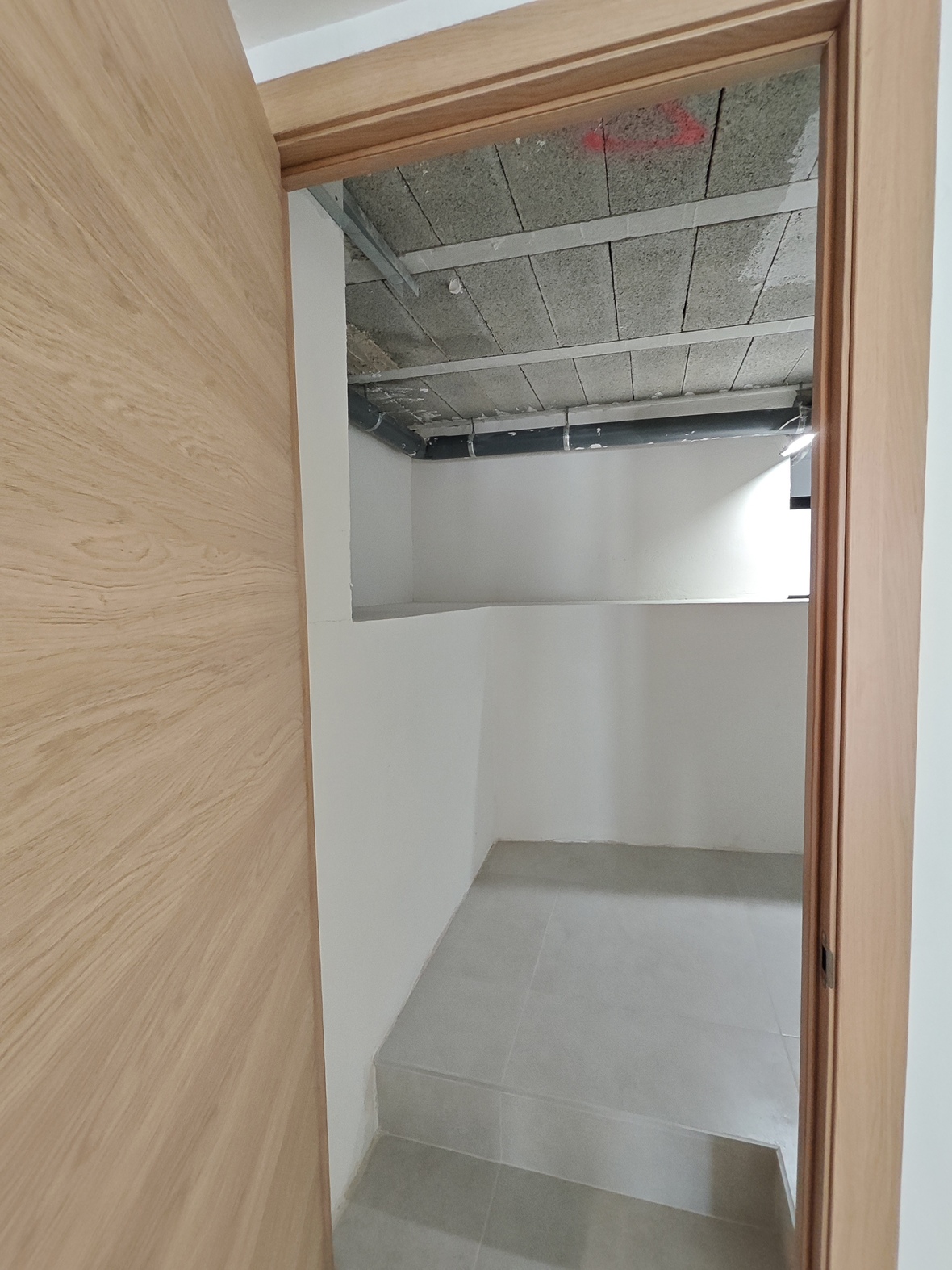 Blick durch eine helle Holztür in einen Abstellraum mit grauen Bodenfliesen, weißen Wänden und sichtbarer Deckeninstallation sowie Rohrleitungen.