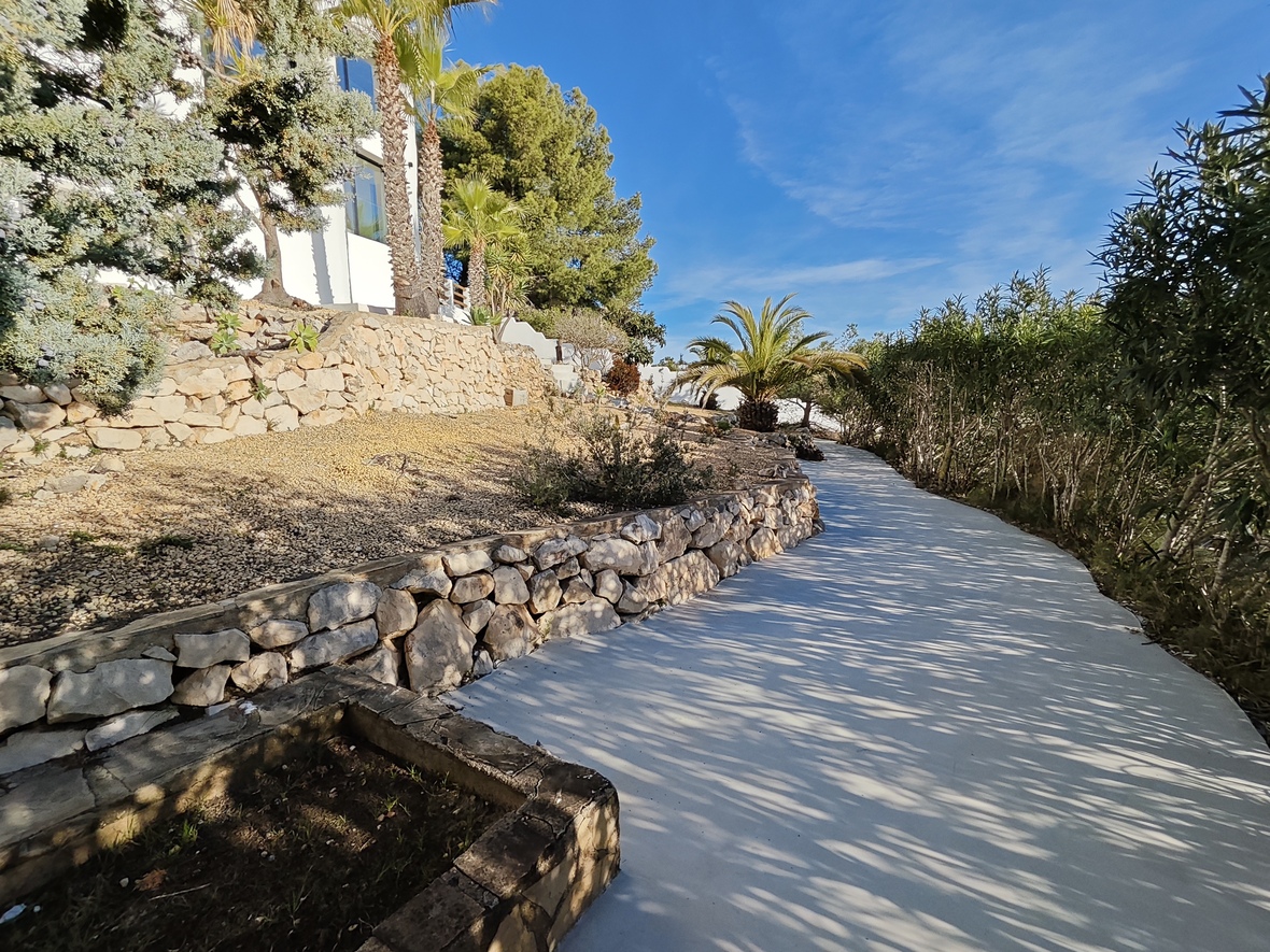 Ein geschwungener, gepflasterter Weg führt durch einen mediterranen Garten mit Trockensteinmauern, Palmen und Pinien vor einer weißen Fassade.