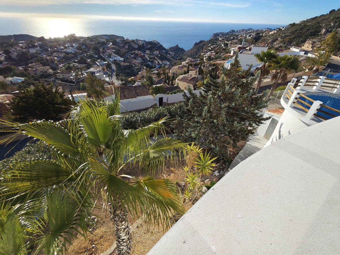 Erhöhter Blick von einer weißen Terrasse auf einen Rundpool mit Holzgeländer, mediterrane Gärten und das weite Meer im Sonnenlicht.