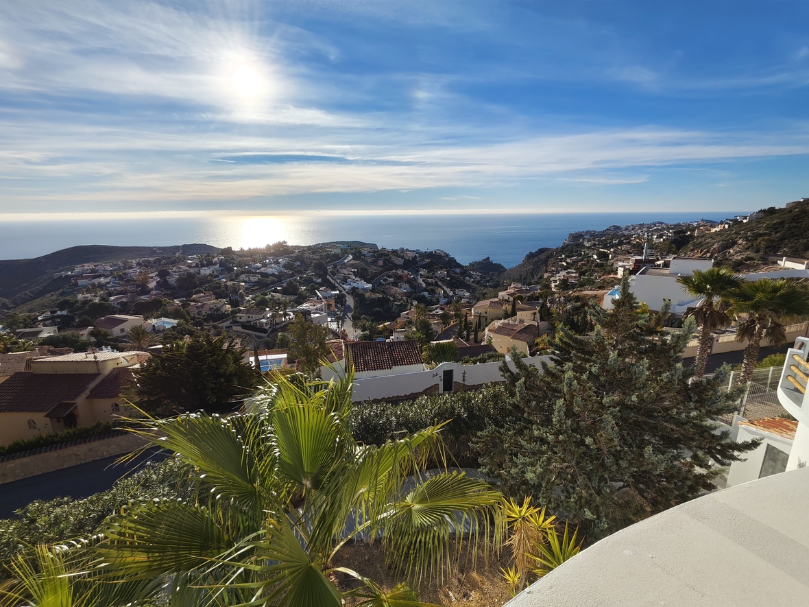 Erhöhter Panoramablick auf ein Küstenwohngebiet und das Mittelmeer, mit weißen Villen, Terrakottadächern und üppigen Palmen unter strahlender Sonne.