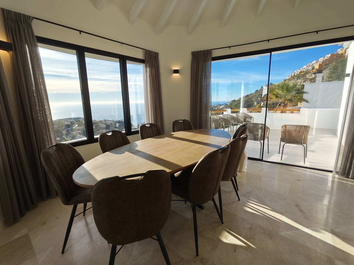 Essbereich mit ovalem Holztisch und Marmorboden, der durch große Fensterfronten einen Panoramablick auf das Meer und direkten Zugang zur Terrasse bietet.