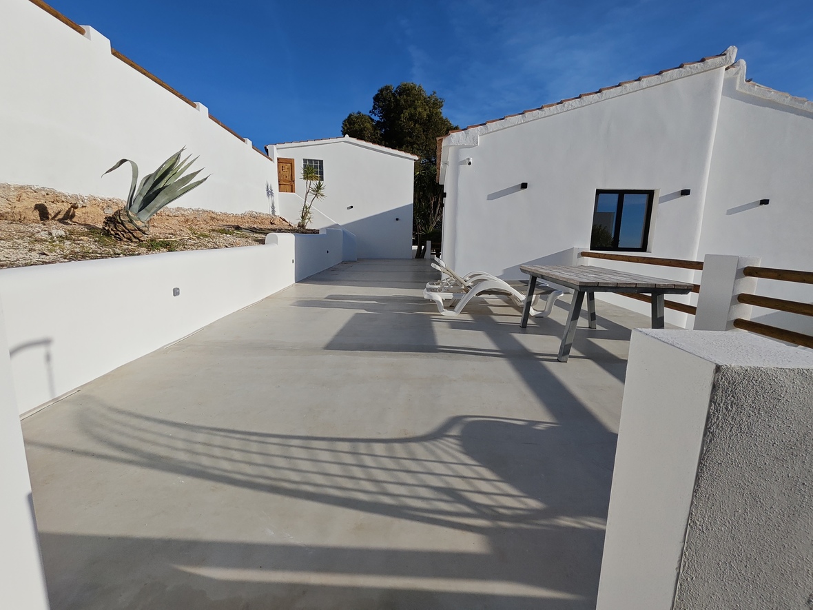 Großzügige Außenterrasse mit poliertem Betonboden, weißen Sonnenliegen und einem Holztisch, umgeben von mediterraner Architektur mit weißen Fassaden.