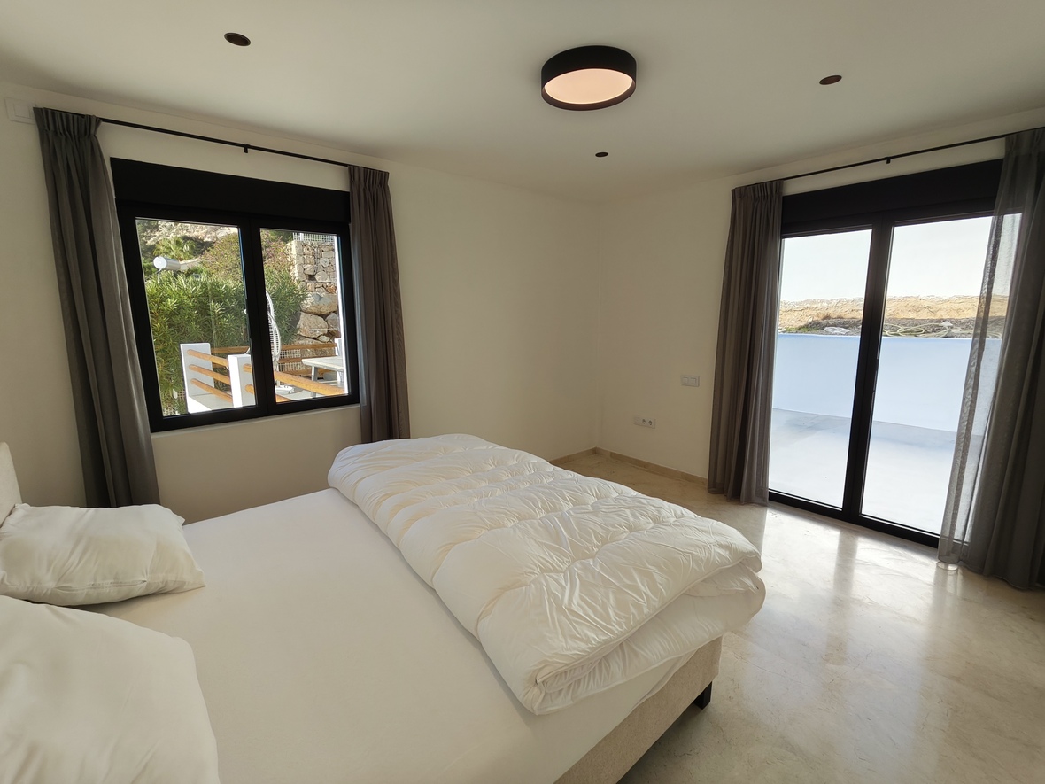 Helles Schlafzimmer mit Marmorboden, schwarz gerahmten Glasschiebetüren zur Terrasse und grauen Vorhängen. Puristische Deckenleuchte und dezente Einbaustrahler.
