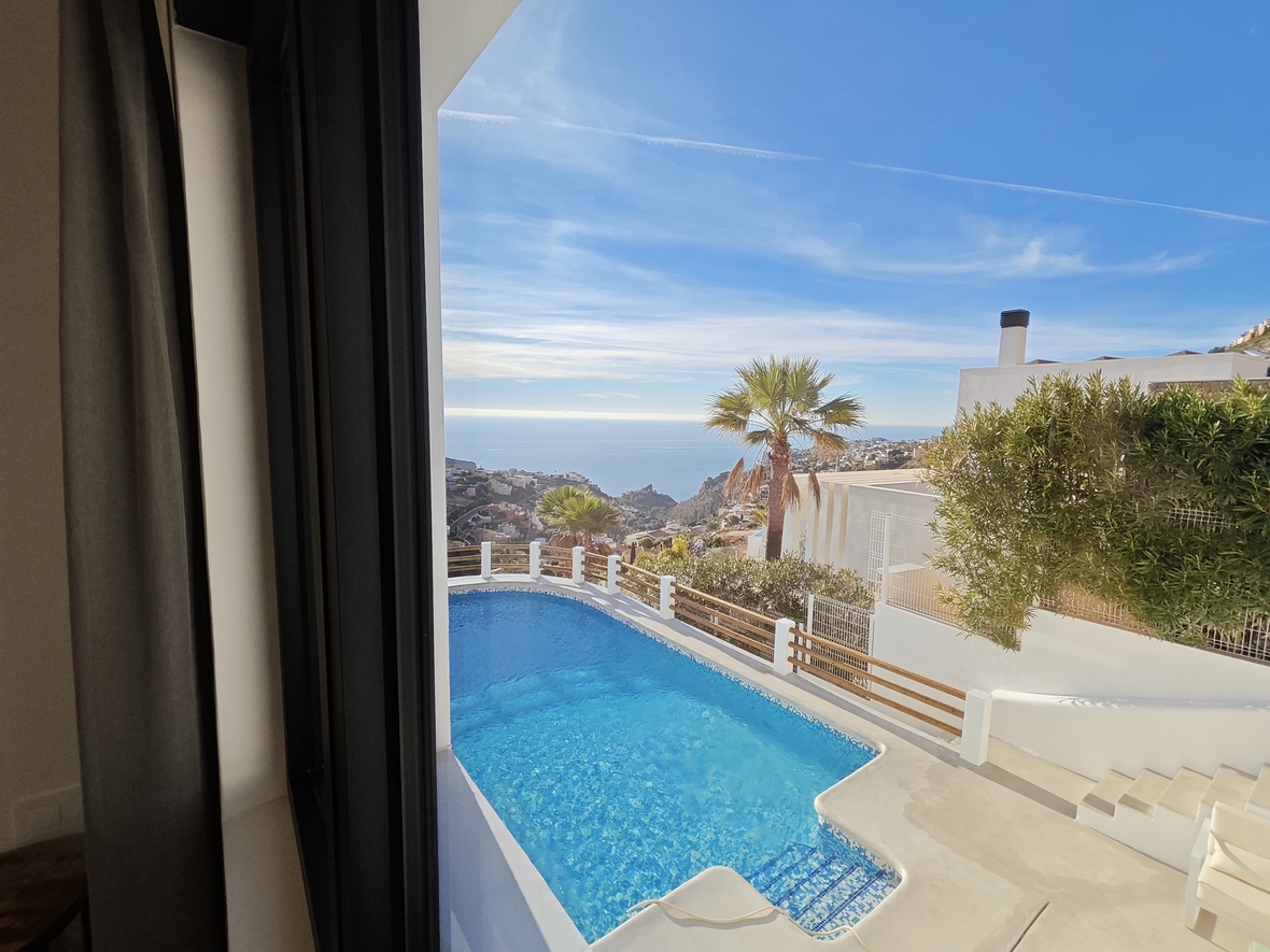 Innenansicht auf einen blauen Mosaikpool und eine weiße Terrasse mit Panoramablick auf die Mittelmeerküste und Palmen unter klarem Himmel.