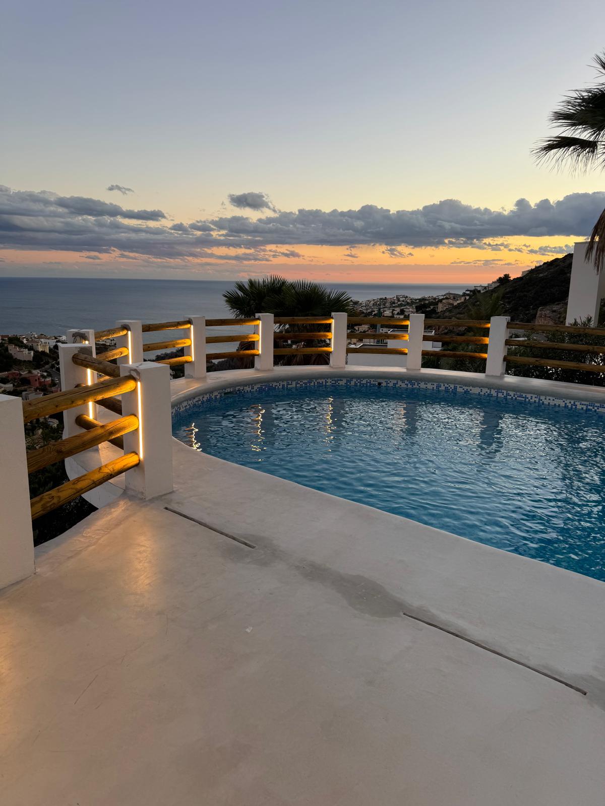 Kreisförmiger Pool mit blauen Mosaikfliesen und LED-Beleuchtung im Holzgeländer, mit Panoramablick auf die Küste während des Sonnenuntergangs.