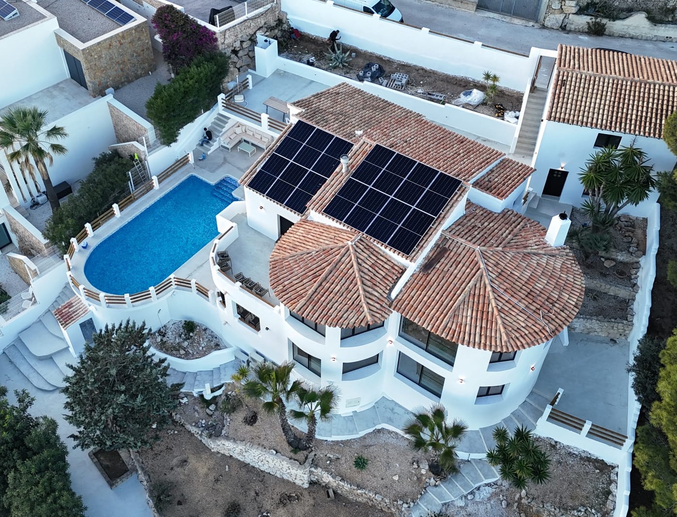 Luftaufnahme einer mediterranen Villa mit Terrakottadächern, Solarmodulen, einem ovalen Pool und geschwungenen Stein-Terrassen auf verschiedenen Ebenen.