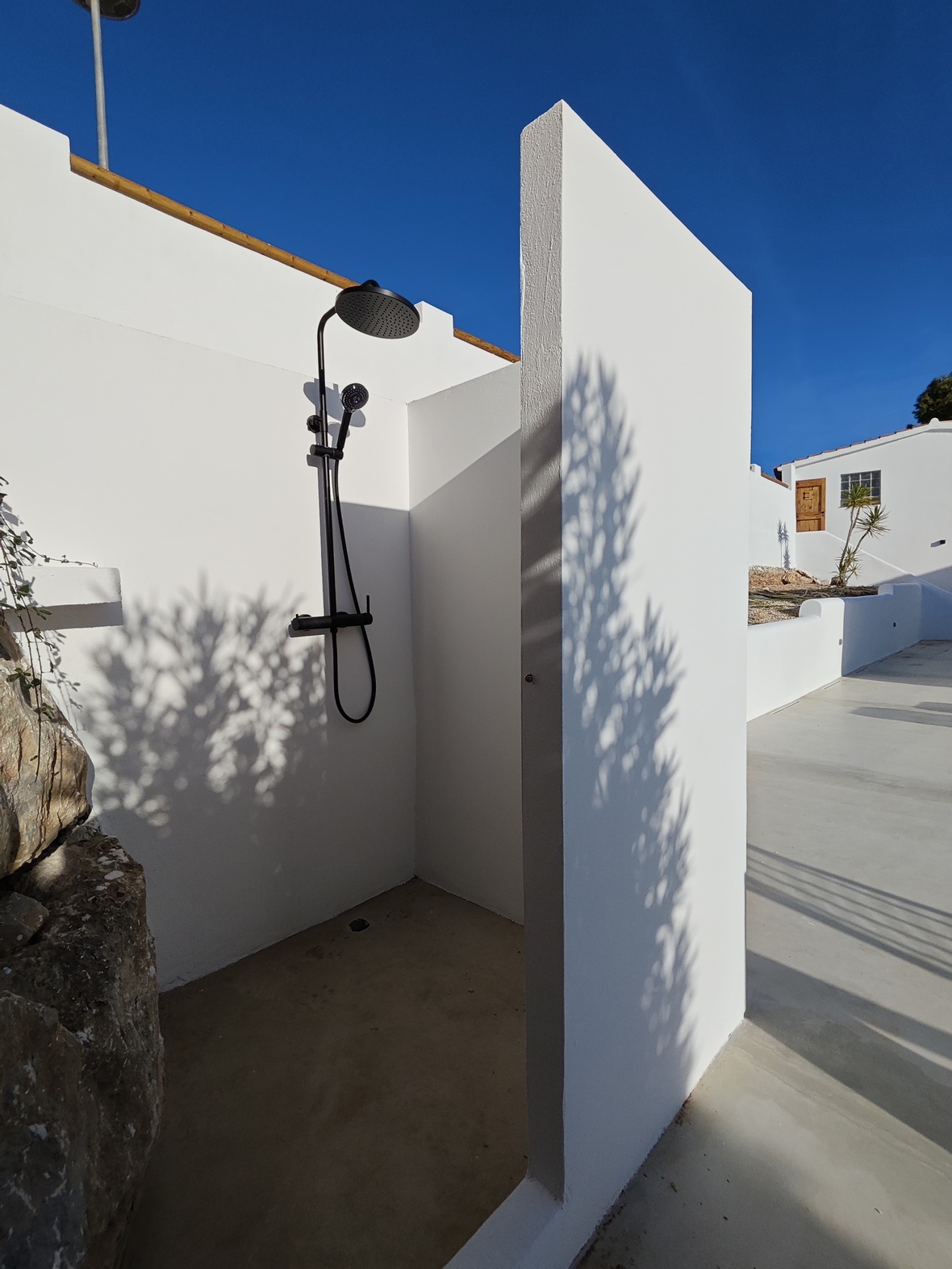 Minimalistische Außendusche mit mattschwarzen Armaturen und weißen Wänden auf einem glatten Betonboden unter strahlend blauem Himmel.