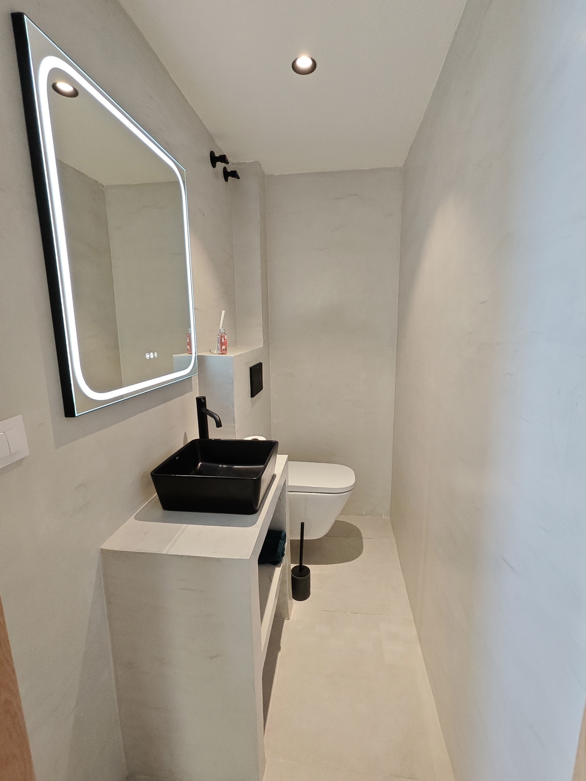 Minimalistisches Badezimmer mit schwarzem Aufsatzwaschbecken, mattschwarzer Armatur, LED-hinterleuchtetem Spiegel und hellgrauen Mikrozementwänden.