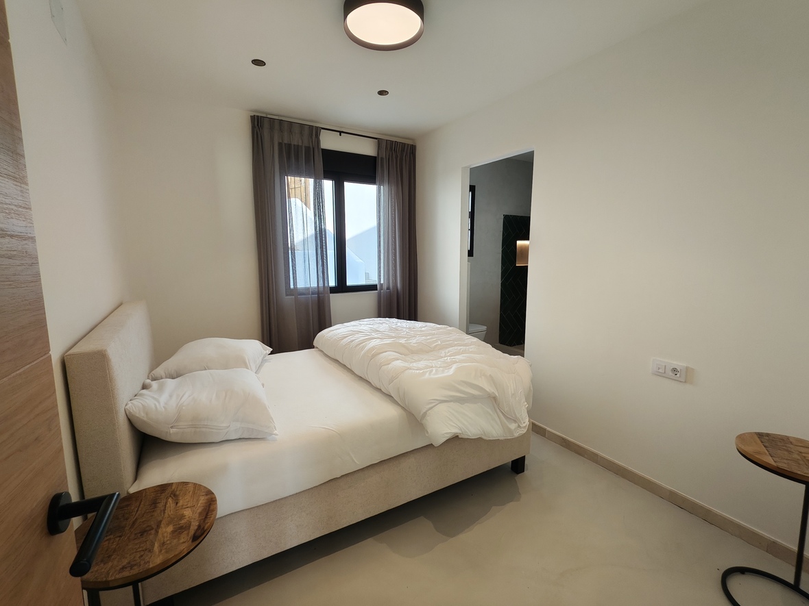 Minimalistisches Schlafzimmer mit gepolstertem Bett, Holznachttischen und angrenzendem Bad mit dunklen Fliesen. Heller Bodenbelag und Fenster mit schwarzen Rahmen.