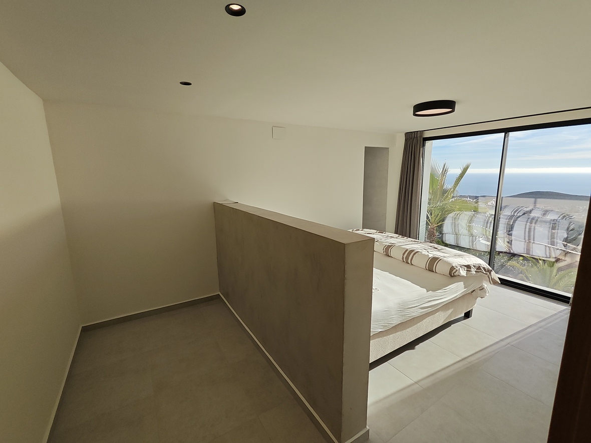 Minimalistisches Schlafzimmer mit halbhoher Trennwand, bodentiefen Glasfronten mit Meerblick und moderner, dezenter Deckenbeleuchtung.