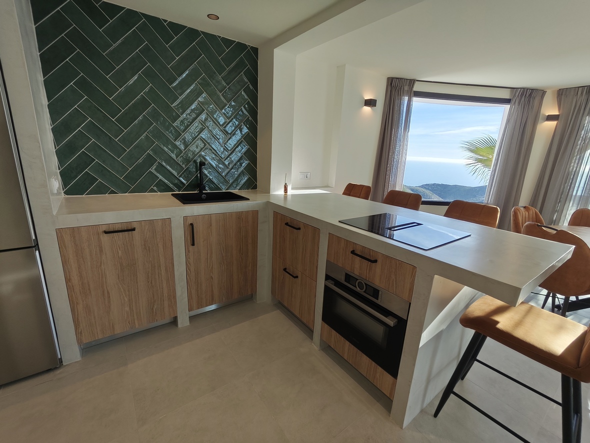Moderne Küche mit grünem Fischgrät-Fliesenspiegel, Holzschränken und einer Kochinsel mit integriertem Kochfeld und Panoramablick auf das Meer.