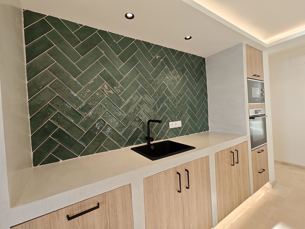 Moderne Küche mit grünem Fliesenspiegel im Fischgrätmuster, schwarzer Spüle und hellen Holzschränken mit integrierten Geräten und LED-Beleuchtung.
