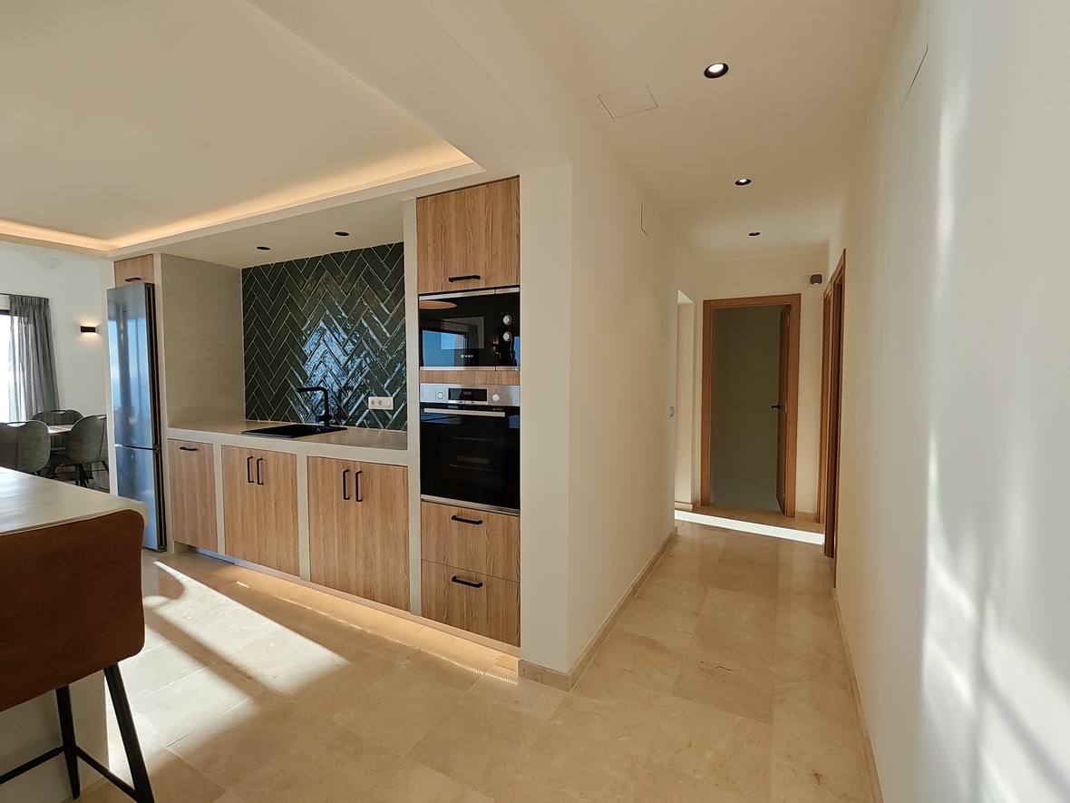 Moderne Küche mit Holzschränken, dunkelgrünem Fischgräten-Fliesenspiegel und Einbaugeräten neben einem hellen Flur mit poliertem Natursteinboden.