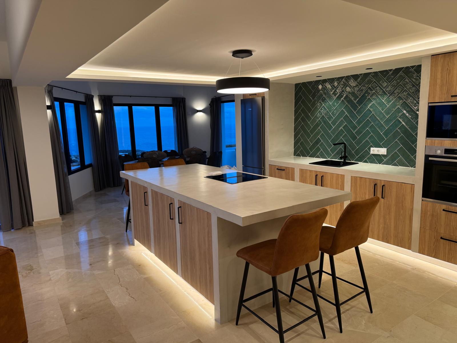 Moderne Küche mit Mikrozement-Insel, grünem Fliesenspiegel im Fischgrätmuster und Panoramablick auf das Meer durch große Fenster zur blauen Stunde.