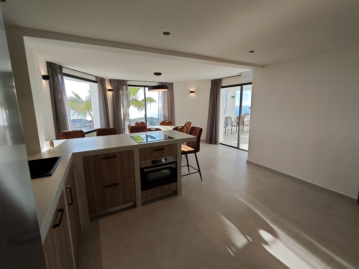 Moderne offene Küche mit Holzfronten, integrierten Geräten und Essbereich, der durch große Fensterfronten einen weiten Blick auf das Meer bietet.
