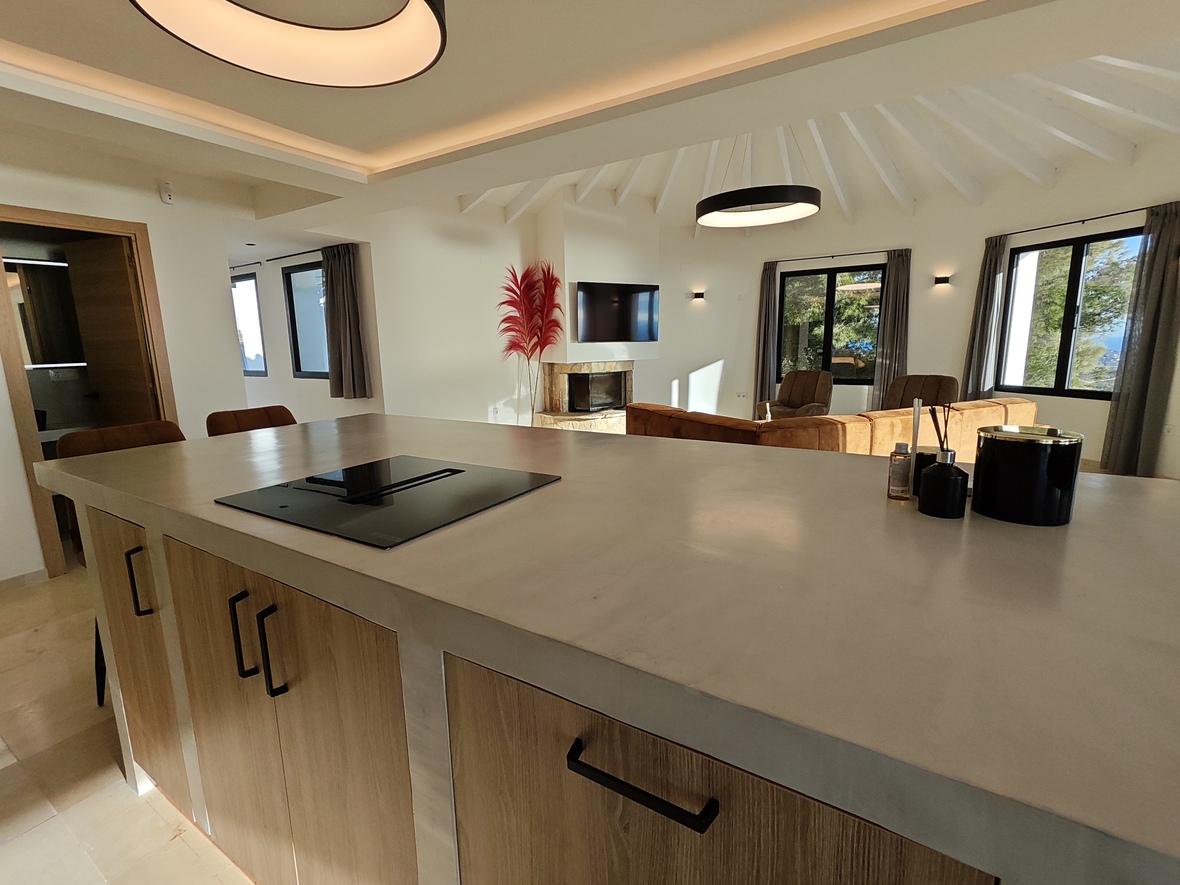 Moderne offene Küche mit Kochinsel, heller Arbeitsplatte und Induktionsfeld, angrenzend an einen Wohnbereich mit weißer Balkendecke und Steinkamin.