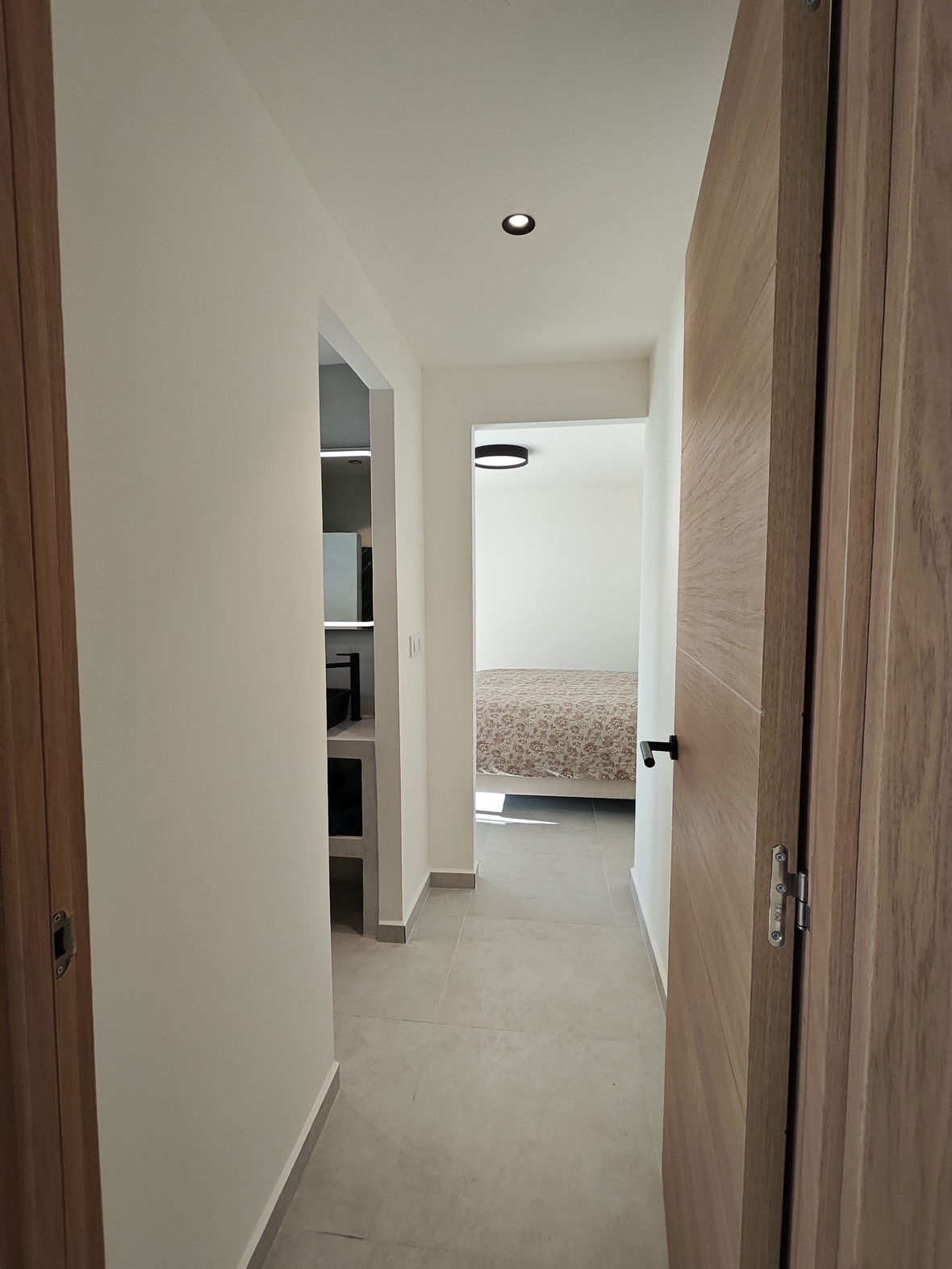 Moderner Flur mit hellen Holztüren und grauen Bodenfliesen, der Zugang zu einem minimalistischen Badezimmer und einem hellen Schlafzimmer bietet.