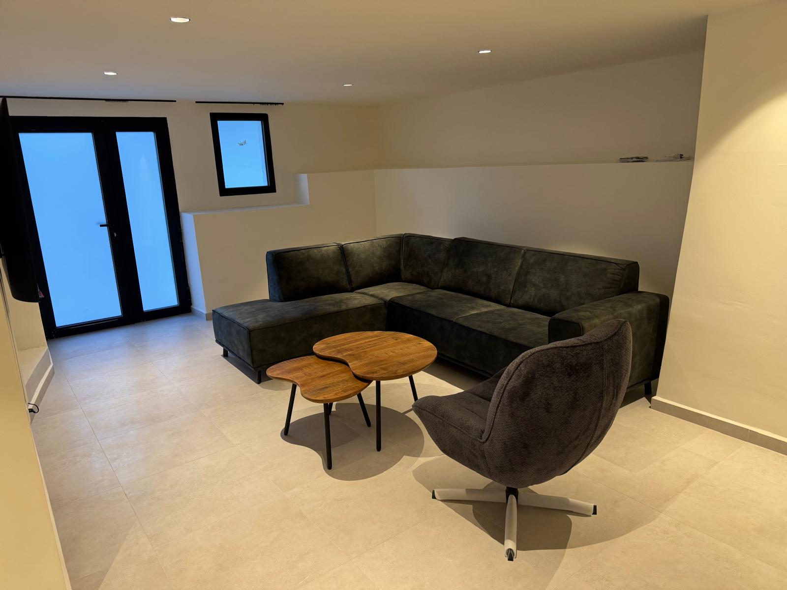 Moderner Wohnbereich mit dunkelgrauem Ecksofa, organisch geformten Couchtischen aus Holz und Drehsessel auf hellen Fliesen mit Deckeneinbaustrahlern.
