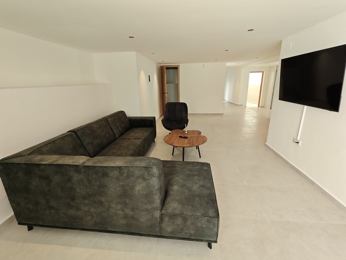 Moderner Wohnbereich mit dunkelgrünem Ecksofa, Couchtisch aus Naturholz und wandmontiertem Flachbildfernseher auf hellem Fliesenboden.