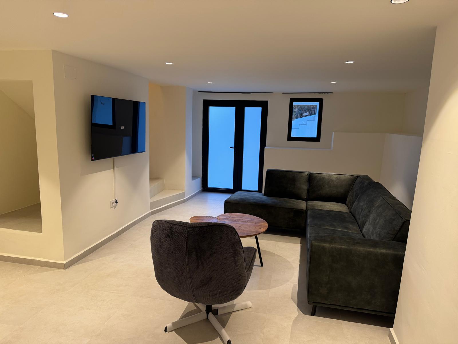 Moderner Wohnbereich mit dunklem Ecksofa, Drehsessel und Wand-TV. Deckeneinbaustrahler und helle Bodenfliesen unterstreichen das minimalistische Ambiente.
