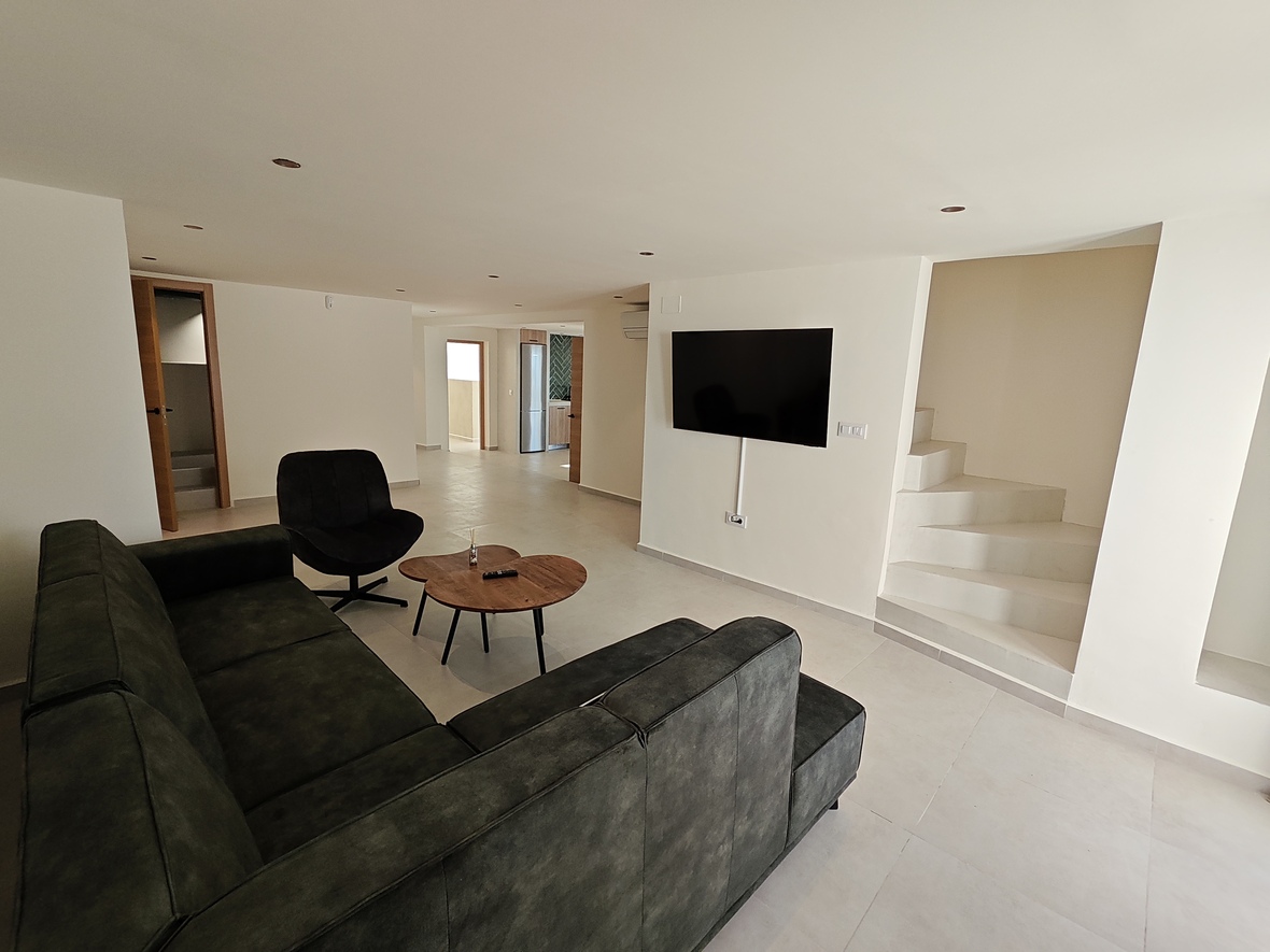 Moderner Wohnbereich mit dunklem Ecksofa, Holz-Couchtisch und Wand-TV neben einer minimalistischen weißen Treppe mit Deckeneinbaustrahlern.