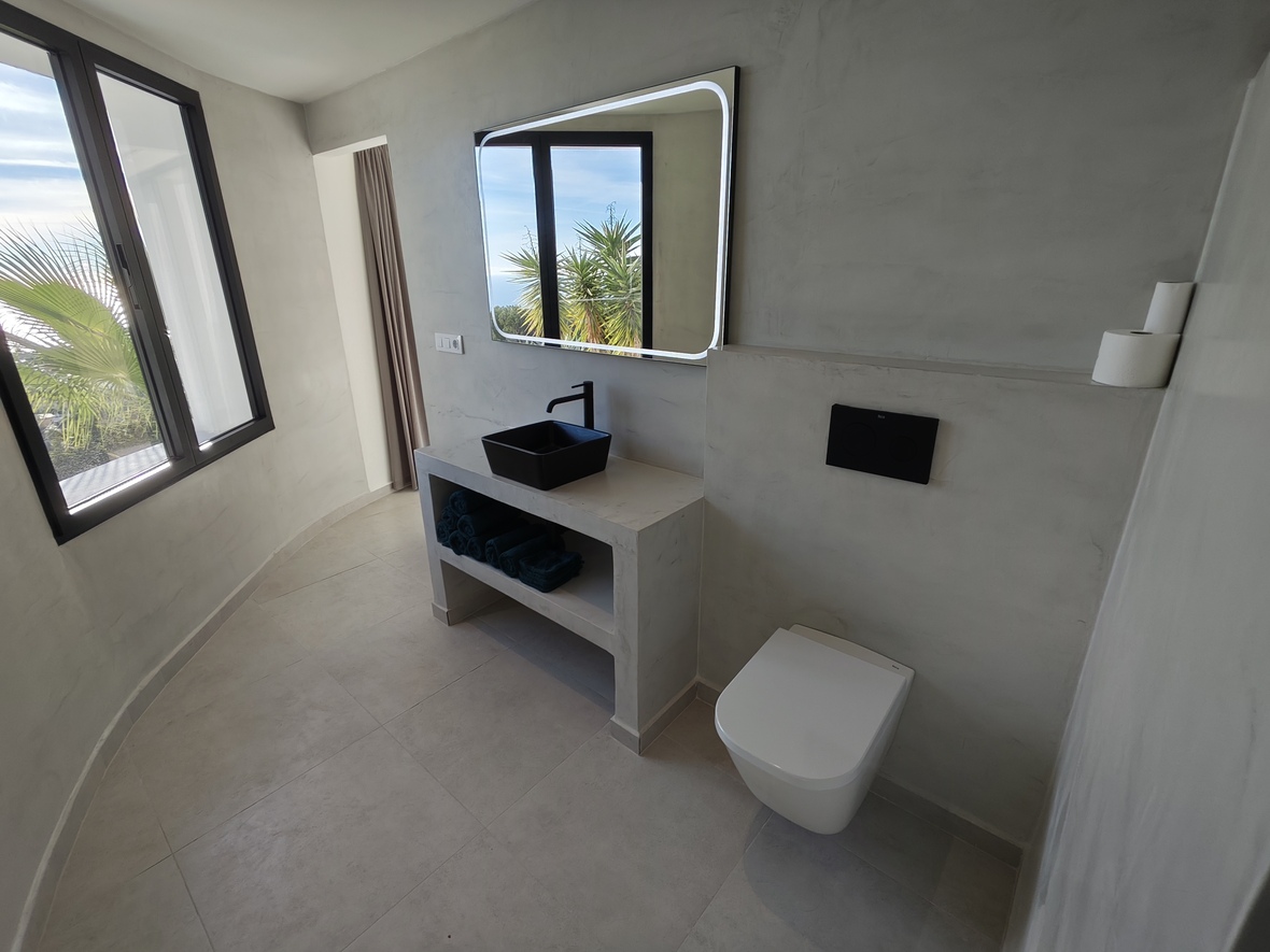 Modernes Badezimmer mit Mikrozementwänden, schwarzem Aufsatzwaschbecken und hinterleuchtetem Spiegel. Große Fenster bieten Tageslicht und Blick auf Palmen.