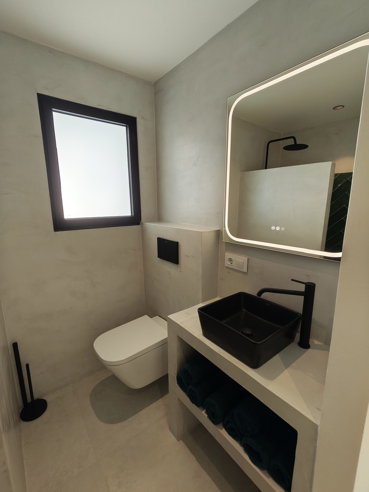 Modernes Badezimmer mit Mikrozementwänden, schwarzem Aufsatzwaschbecken und LED-Spiegel. Ausgestattet mit mattschwarzen Armaturen und einem Wand-WC.