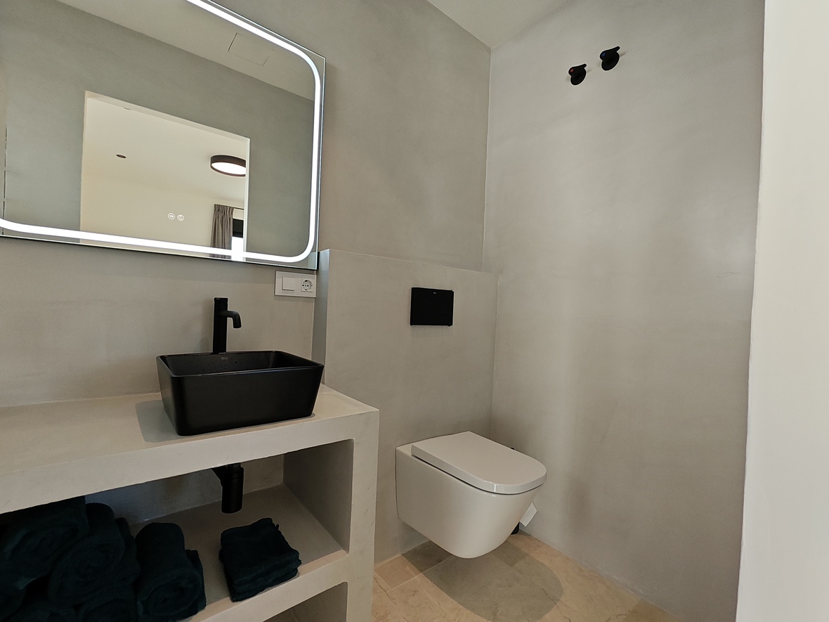 Modernes Gäste-WC mit schwarzem Aufsatzwaschbecken, hintergrundbeleuchtetem Spiegel und wandhängendem WC vor grauen Mikrozementwänden.
