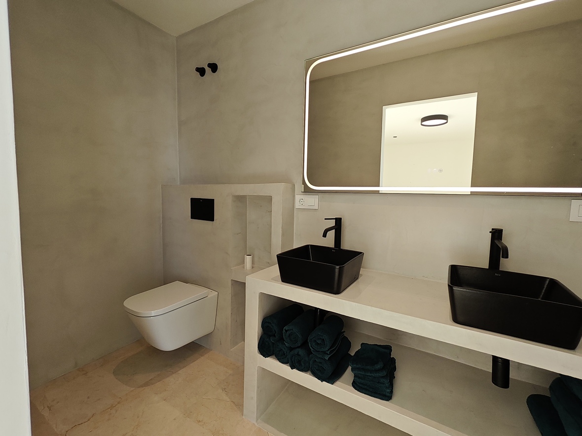 Modernes, minimalistisches Badezimmer mit grauen Mikrozementwänden, Doppelwaschbecken mit schwarzen Schalen und einem großen LED-Lichtspiegel.