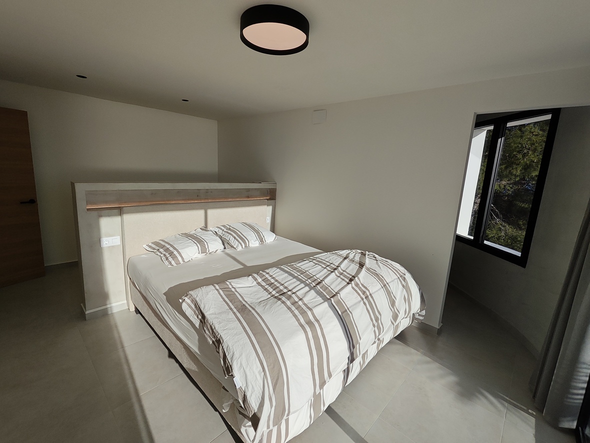 Modernes Schlafzimmer mit integriertem Trennwand-Kopfteil und LED-Beleuchtung, großformatigen Fliesen und einem schwarz gerahmten Fenster mit Blick ins Grüne.