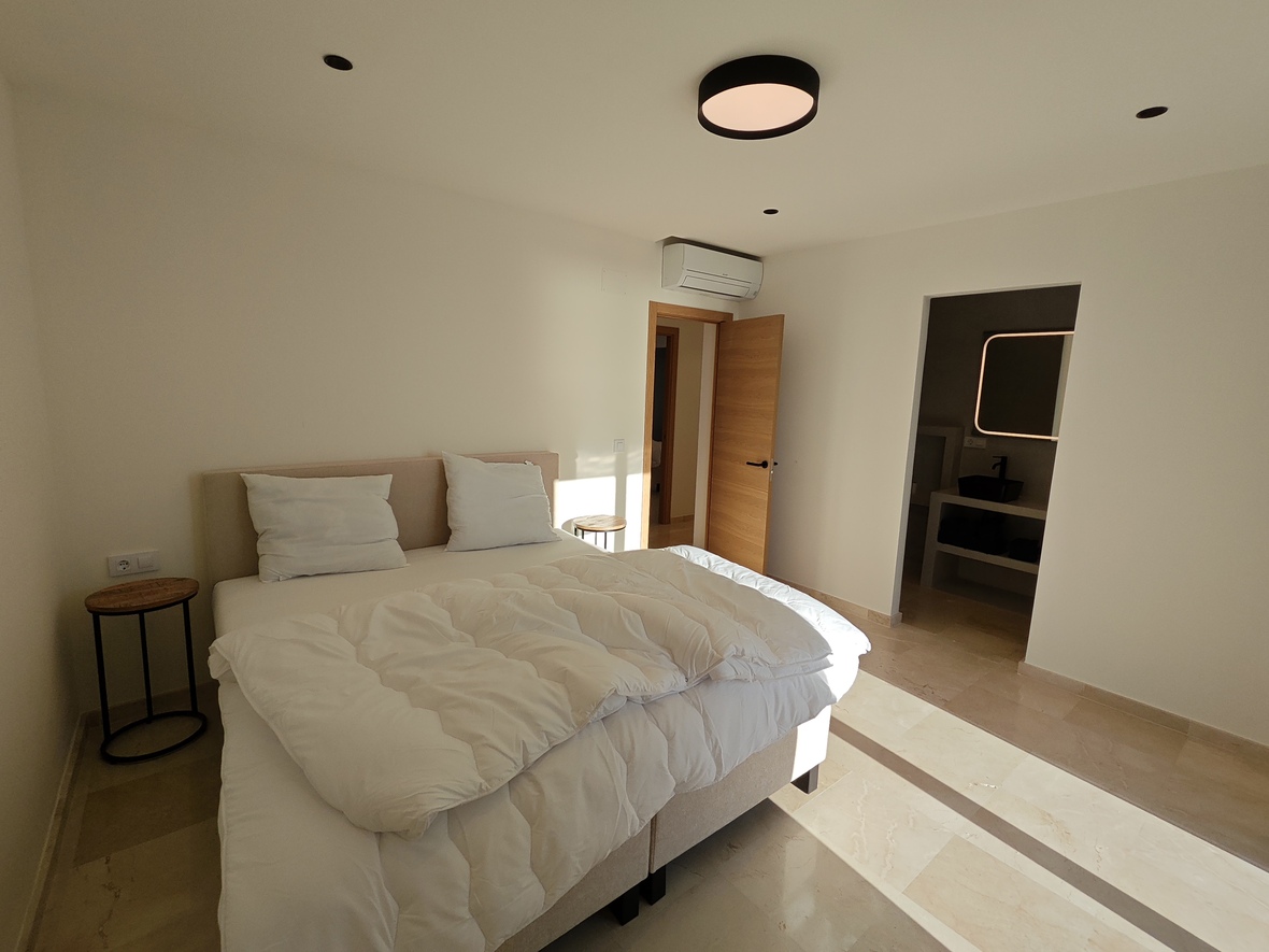 Modernes Schlafzimmer mit Steinfliesenboden, gepolstertem beigem Kopfteil und minimalistischen Nachttischen. Inklusive Klimaanlage und En-Suite-Bad mit hinterleuchtetem Spiegel.