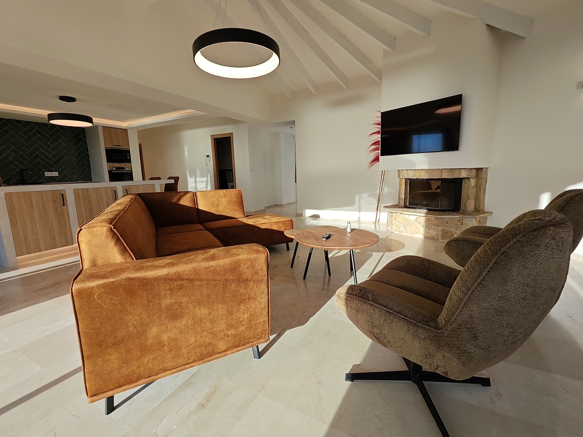 Modernes Wohnzimmer mit ockerfarbenem Samtsofa, Natursteinkamin und Sichtbalkendecke. Im Hintergrund die offene Küche mit grünem Fliesenspiegel und hellen Holzfronten.