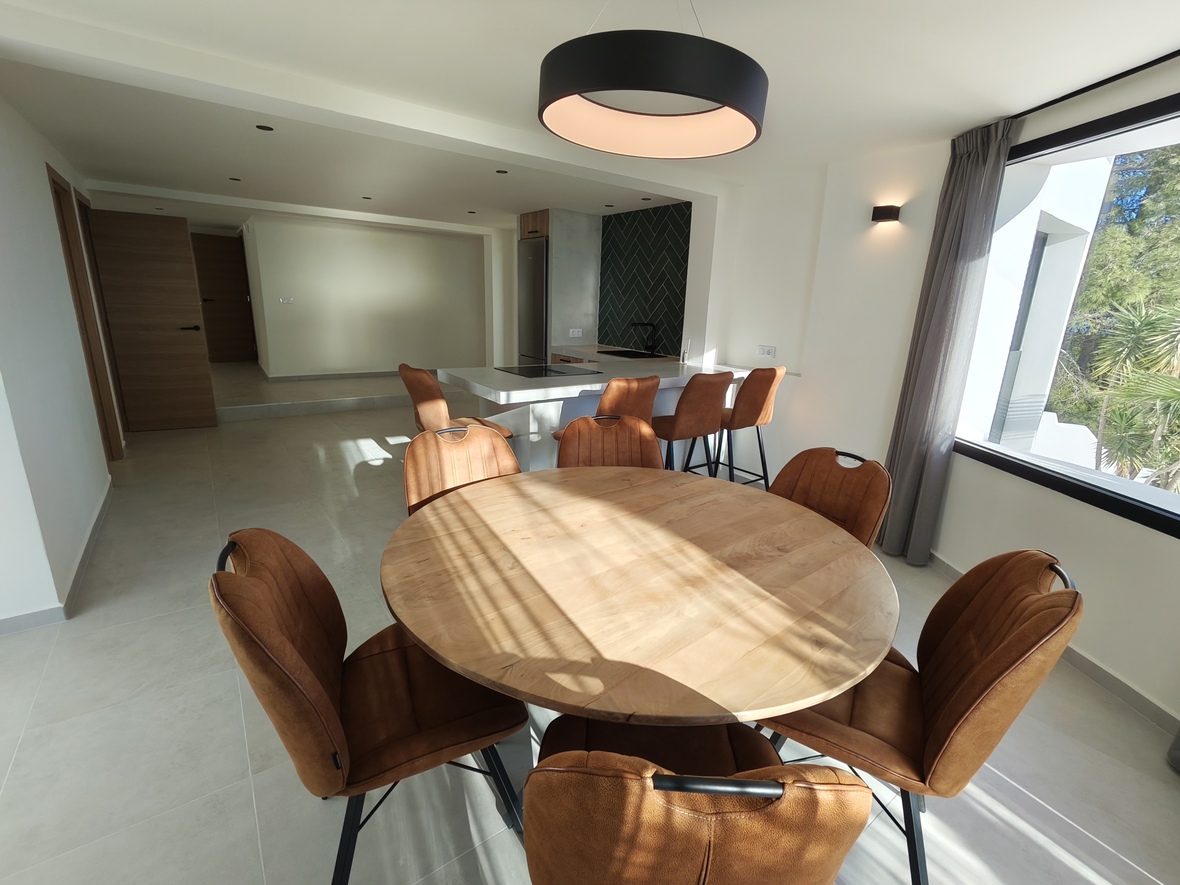 Offener Essbereich mit rundem Holztisch, cognacfarbenen Polsterstühlen und Ringpendelleuchte, angrenzend an eine moderne Küche mit weißer Kochinsel.