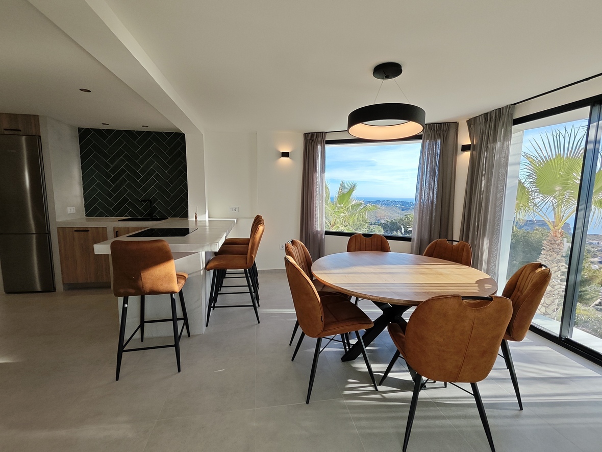 Offener Essbereich mit rundem Holztisch, cognacfarbenen Stühlen und moderner Küche mit Fischgrät-Fliesenspiegel und Panoramablick auf das Meer.