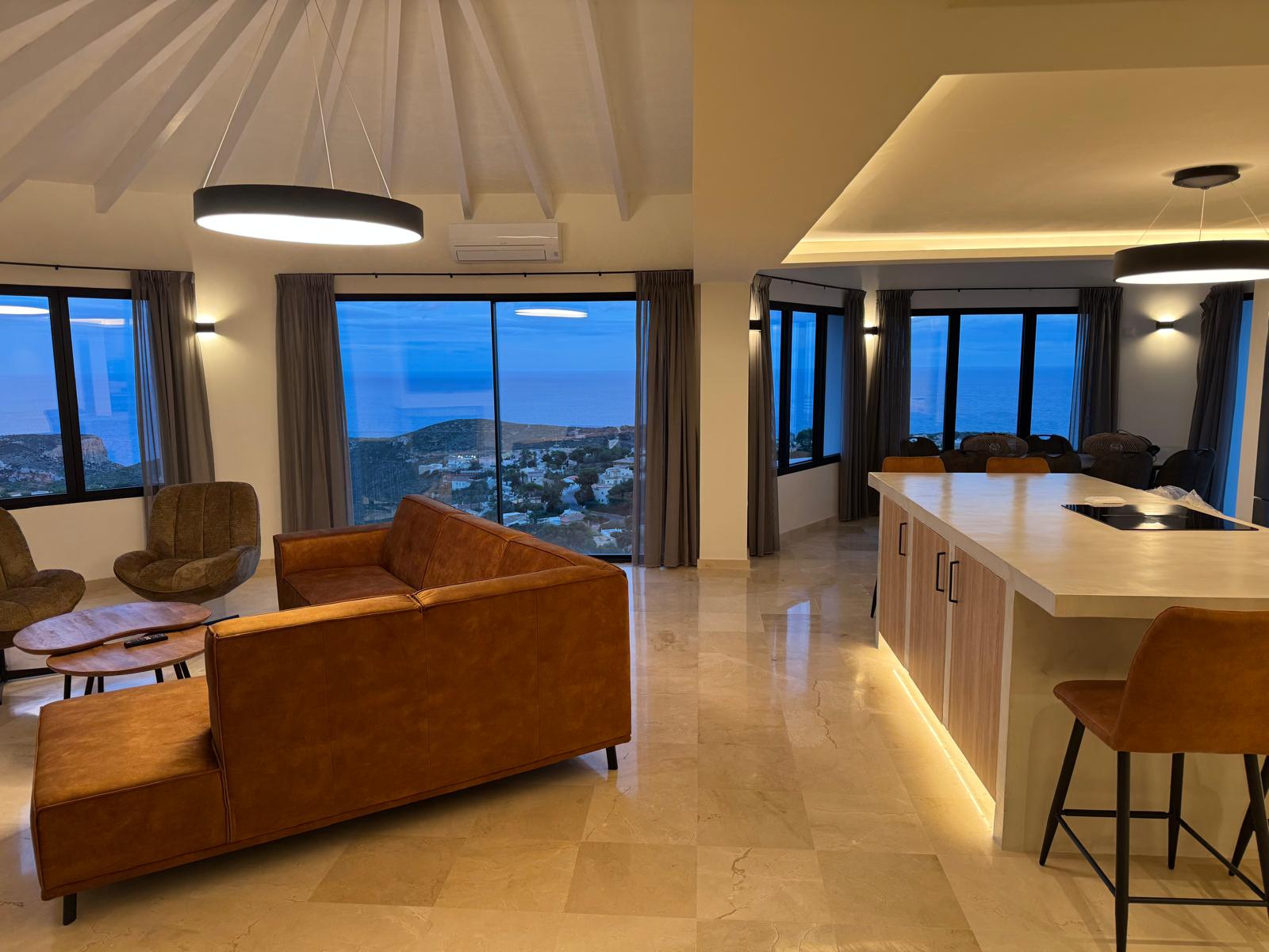 Offener Wohnbereich mit cognacfarbenem Ledersofa, Kücheninsel mit LED-Beleuchtung und großen Fenstern mit Panoramablick auf das Meer bei Dämmerung.