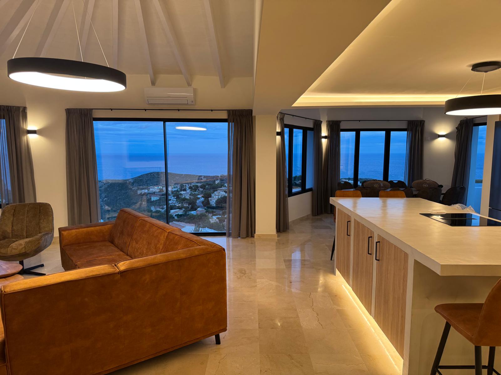 Offener Wohnbereich mit cognacfarbenem Ledersofa und Kücheninsel mit integrierter Beleuchtung, die durch große Fensterfronten den Blick auf das Meer freigibt.