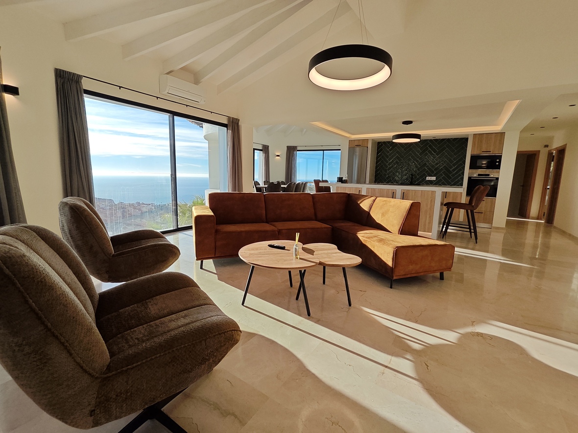 Offener Wohnbereich mit terrakottafarbenem Ecksofa, Sichtbalkendecke und bodentiefen Fenstern mit Meerblick über poliertem Natursteinboden.