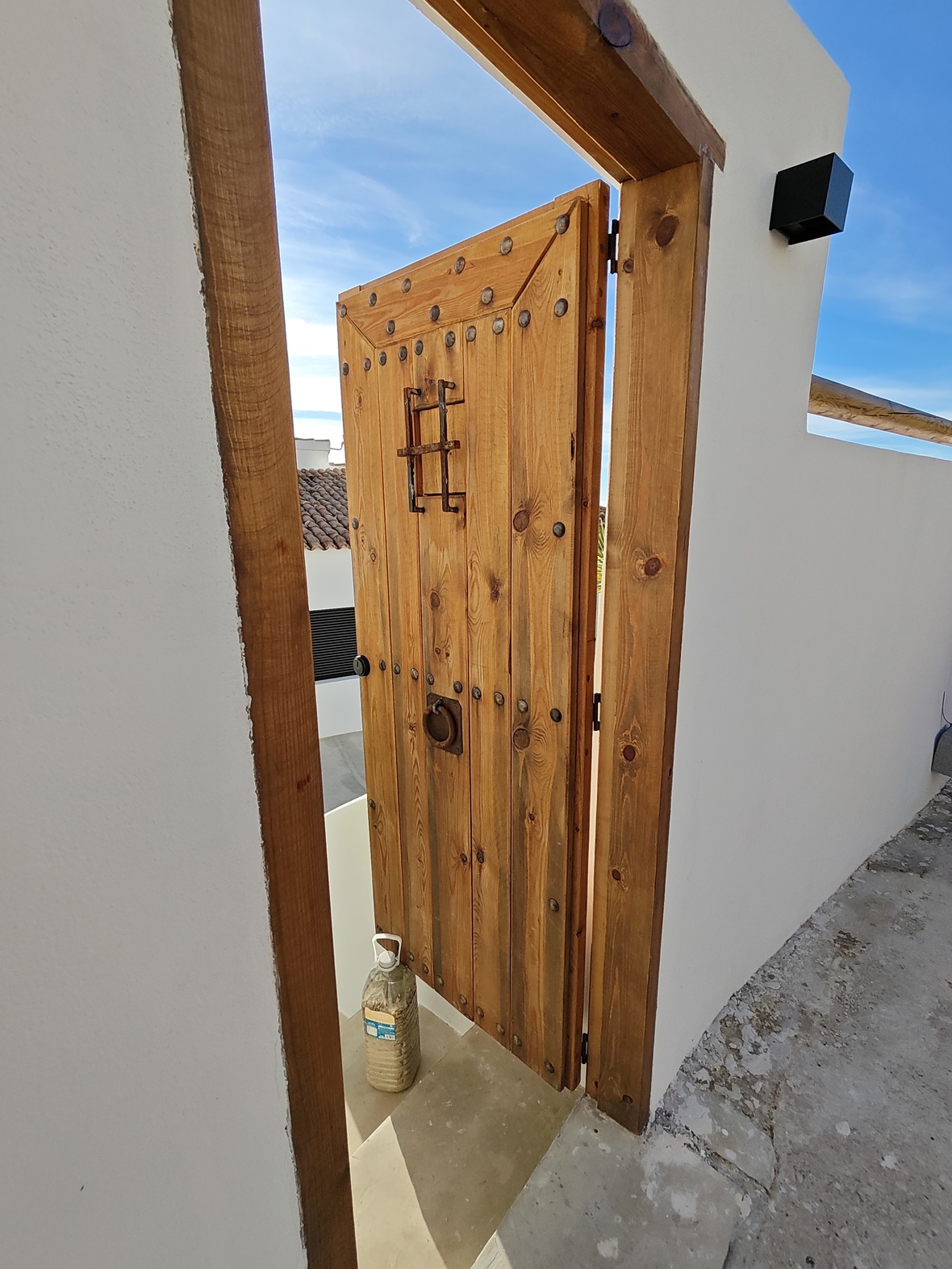 Rustikale Holztür mit Eisenbeschlägen und vergittertem Sichtfenster, eingefasst in einen massiven Holzrahmen vor weißer Fassade.