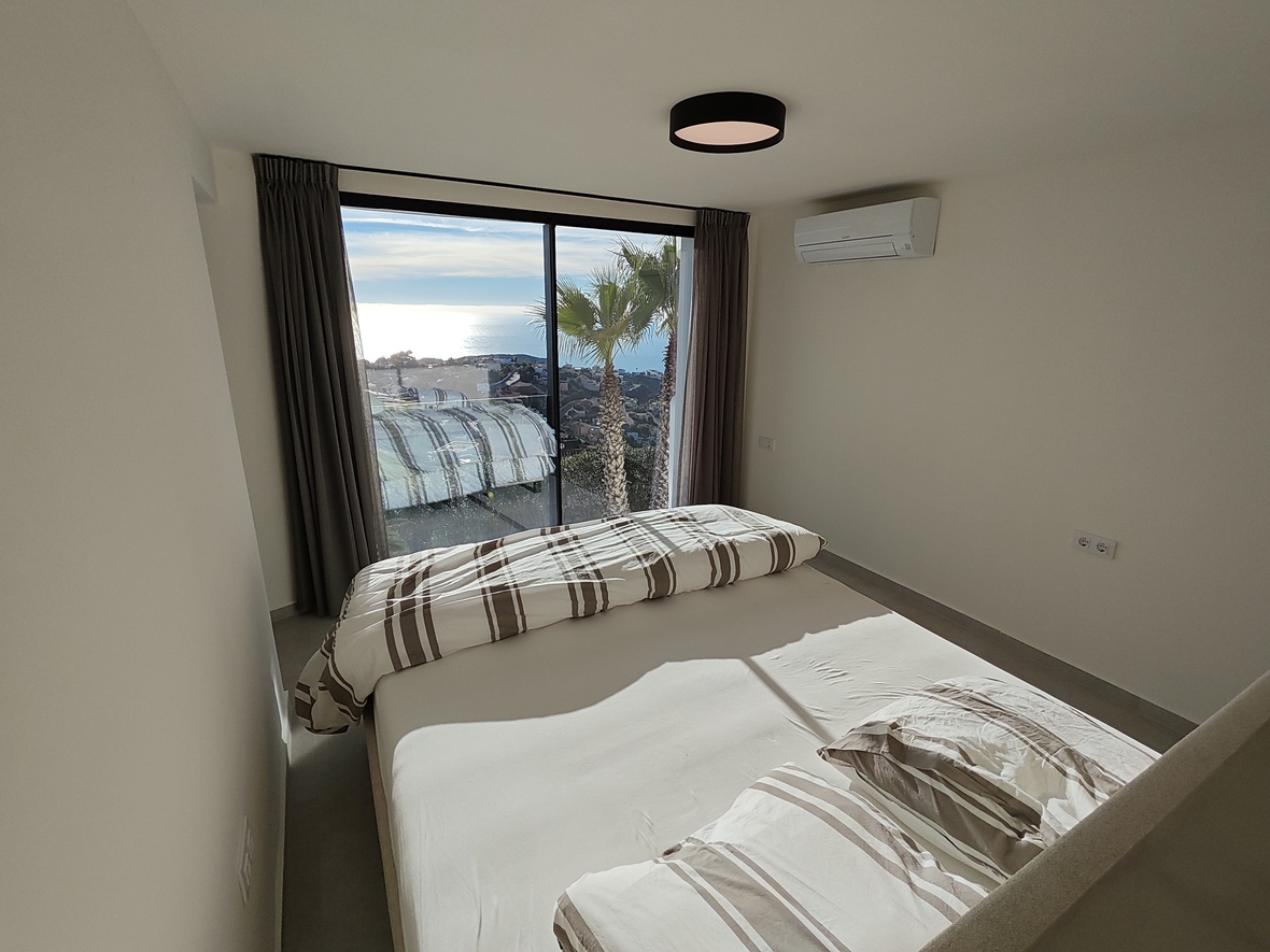 Schlafzimmer mit bodentiefen Glasschiebetüren, Meerblick und Palmen. Ausgestattet mit minimalistischen weißen Wänden, gestreifter Bettwäsche und Wandklimaanlage.