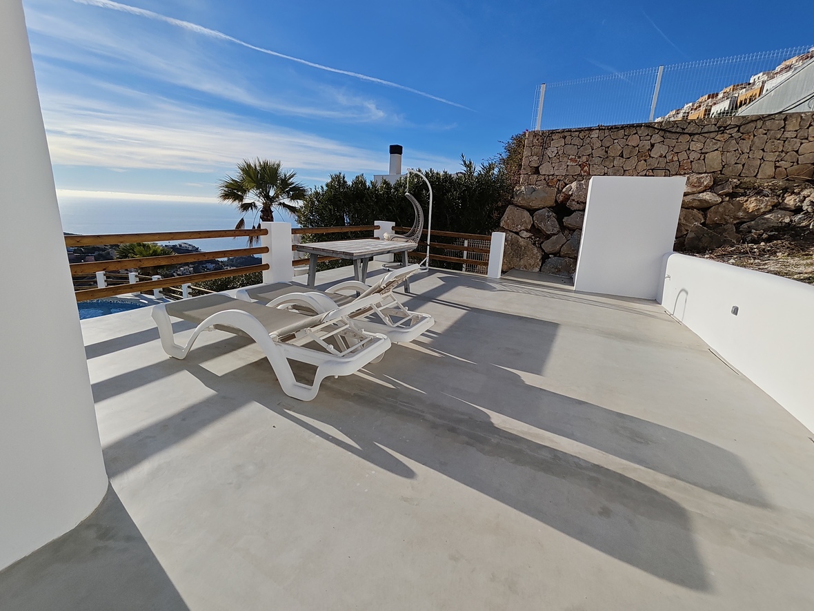 Sonnenterrasse mit weißen Liegen, Holztisch und Hängesessel. Mit Panoramablick auf das Meer, Natursteinmauern und glattem Betonboden.