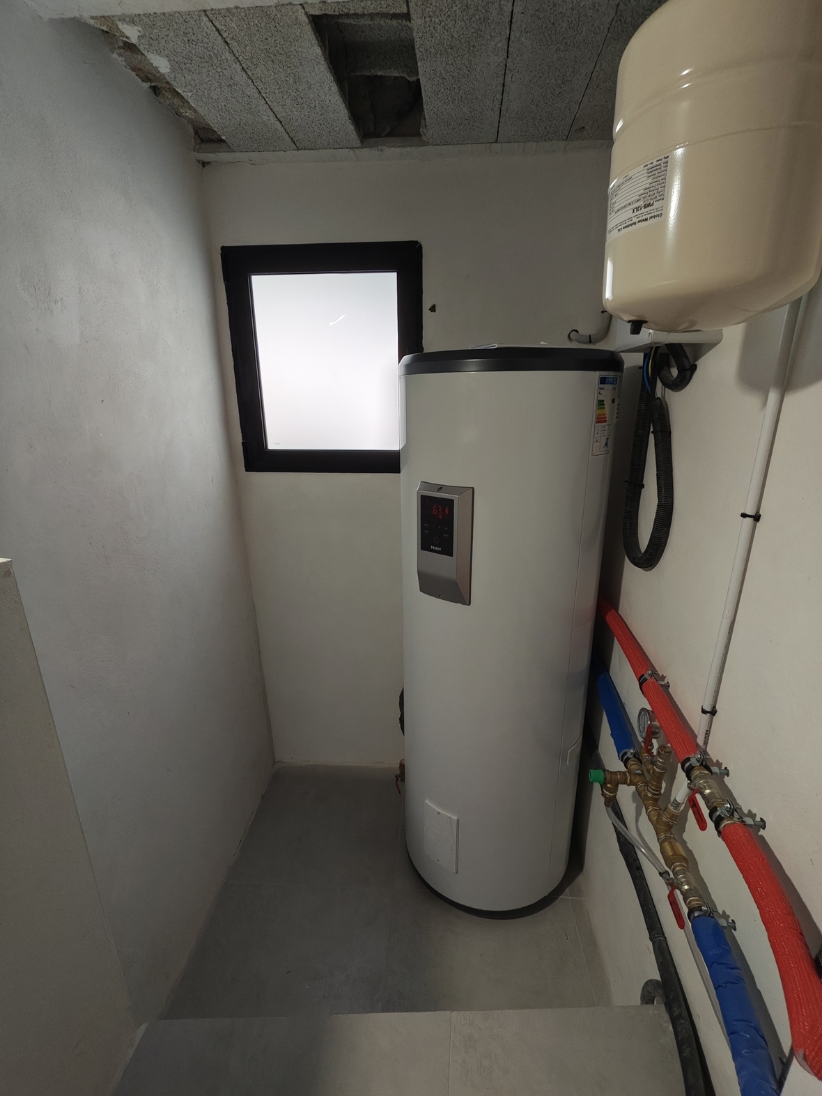 Technikraum mit Haier Warmwasserspeicher, Ausdehnungsgefäß und isolierten Rohrleitungen. Ausgestattet mit einem Milchglasfenster und grauen Bodenfliesen.