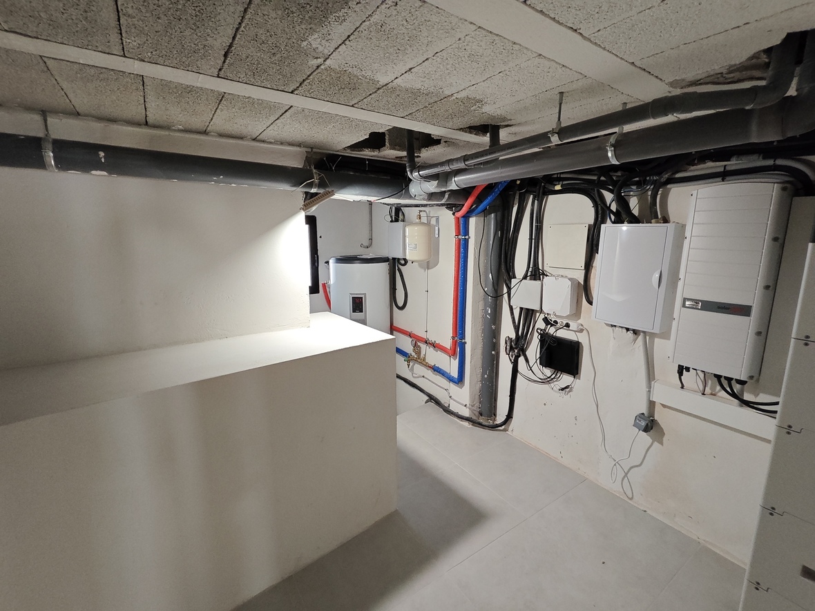 Technikraum mit SolarEdge-Wechselrichter, Elektroverteilerkasten und Warmwassersystem mit sichtbaren PEX-Rohren und Betonsteindecke.