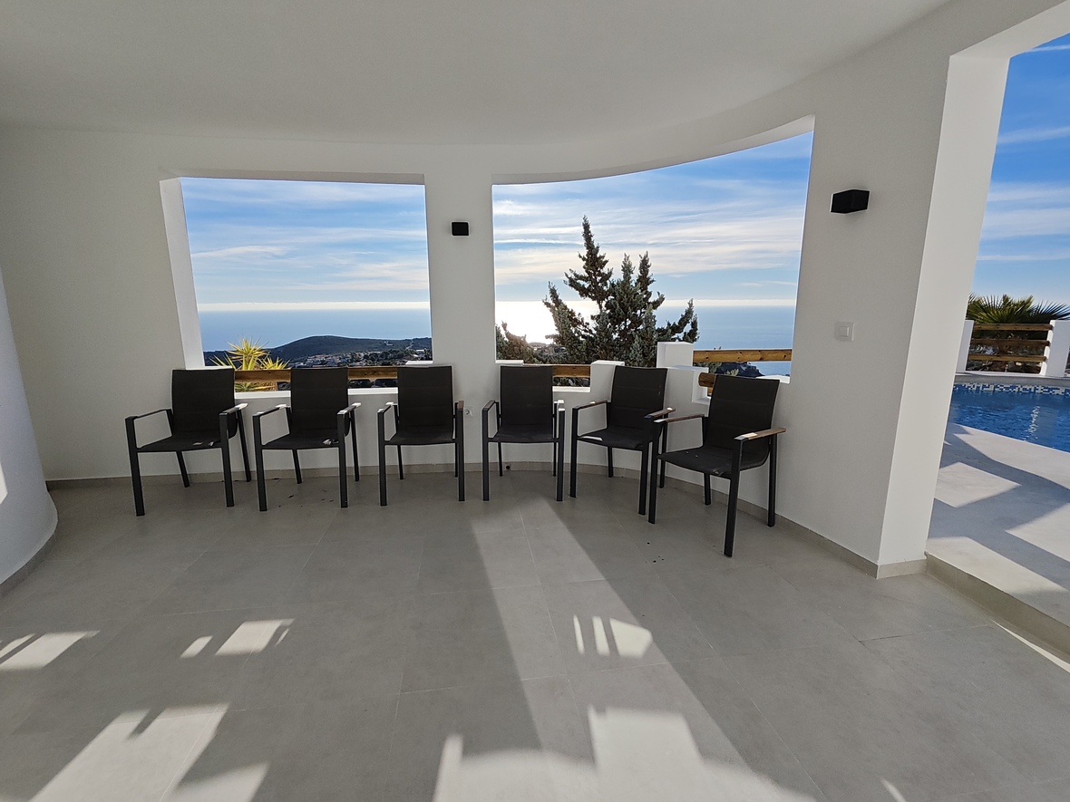 Überdachte Terrasse mit modernen schwarzen Sesseln, hellgrauem Fliesenboden und großen Maueröffnungen mit Panoramablick auf das Meer und den angrenzenden Pool.