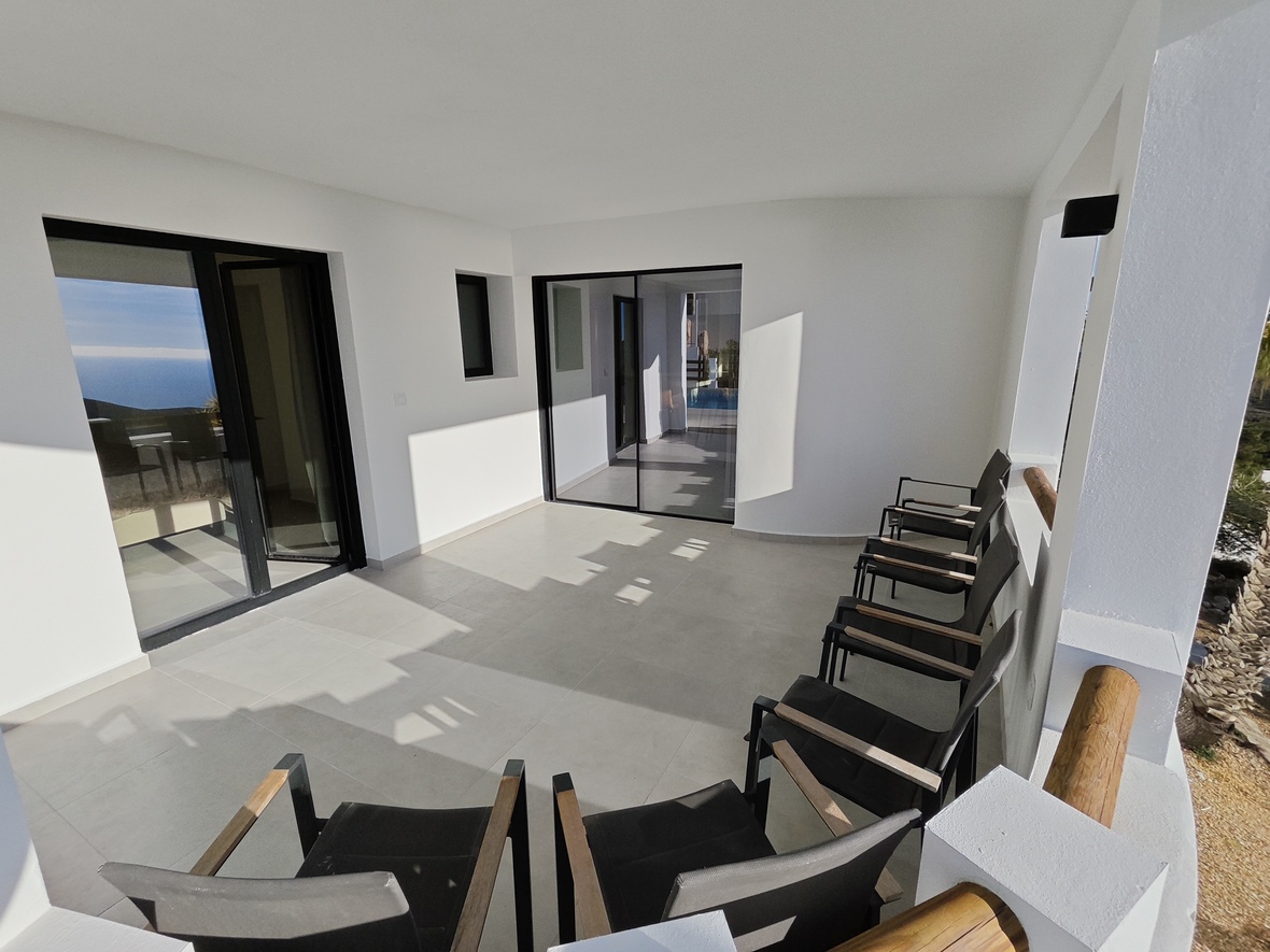 Überdachte Terrasse mit weißen Wänden, hellgrauen Fliesen und schwarzen Schiebetüren. Moderne schwarze Sessel mit Holzarmlehnen bieten Blick auf das Meer.