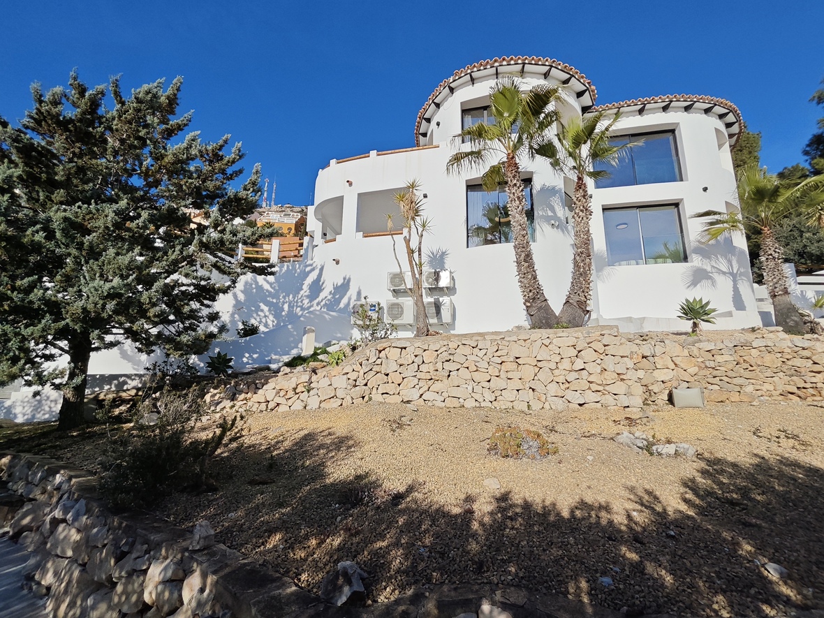 Weiße mediterrane Villa mit runden Bauelementen, Natursteinmauern und einer Gartenanlage mit Palmen auf einem terrassierten Grundstück.