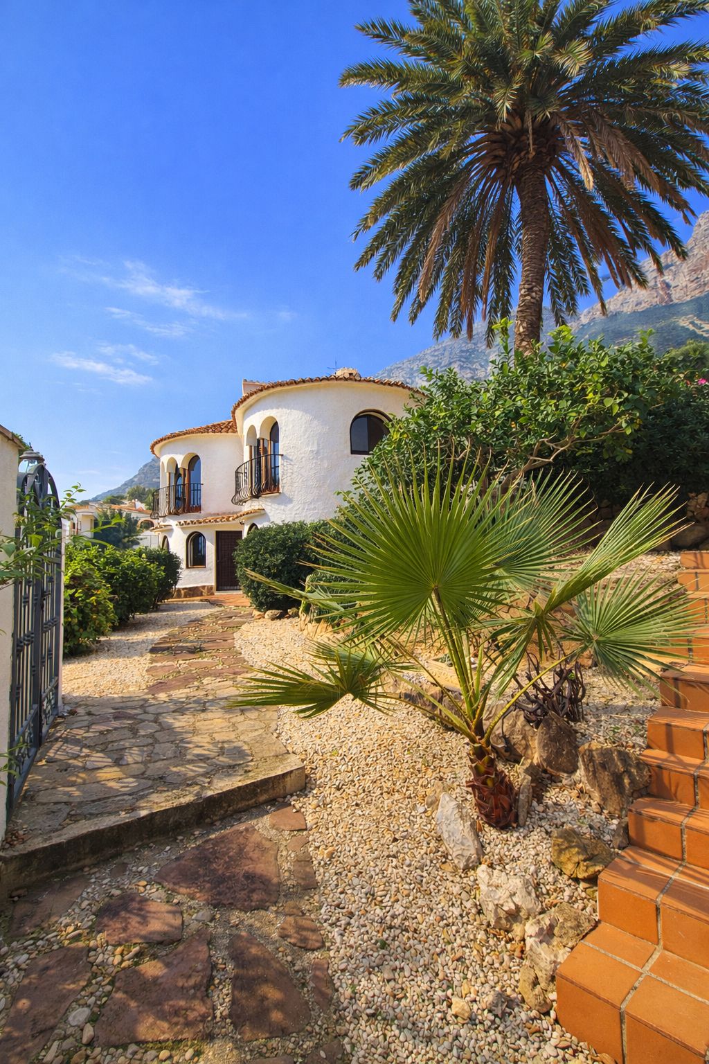 Außenansicht einer weißen mediterranen Villa mit Rundtürmen, Rundbogenfenstern und einem Steinpfad in einem Garten mit Palmen vor einer Bergkulisse.