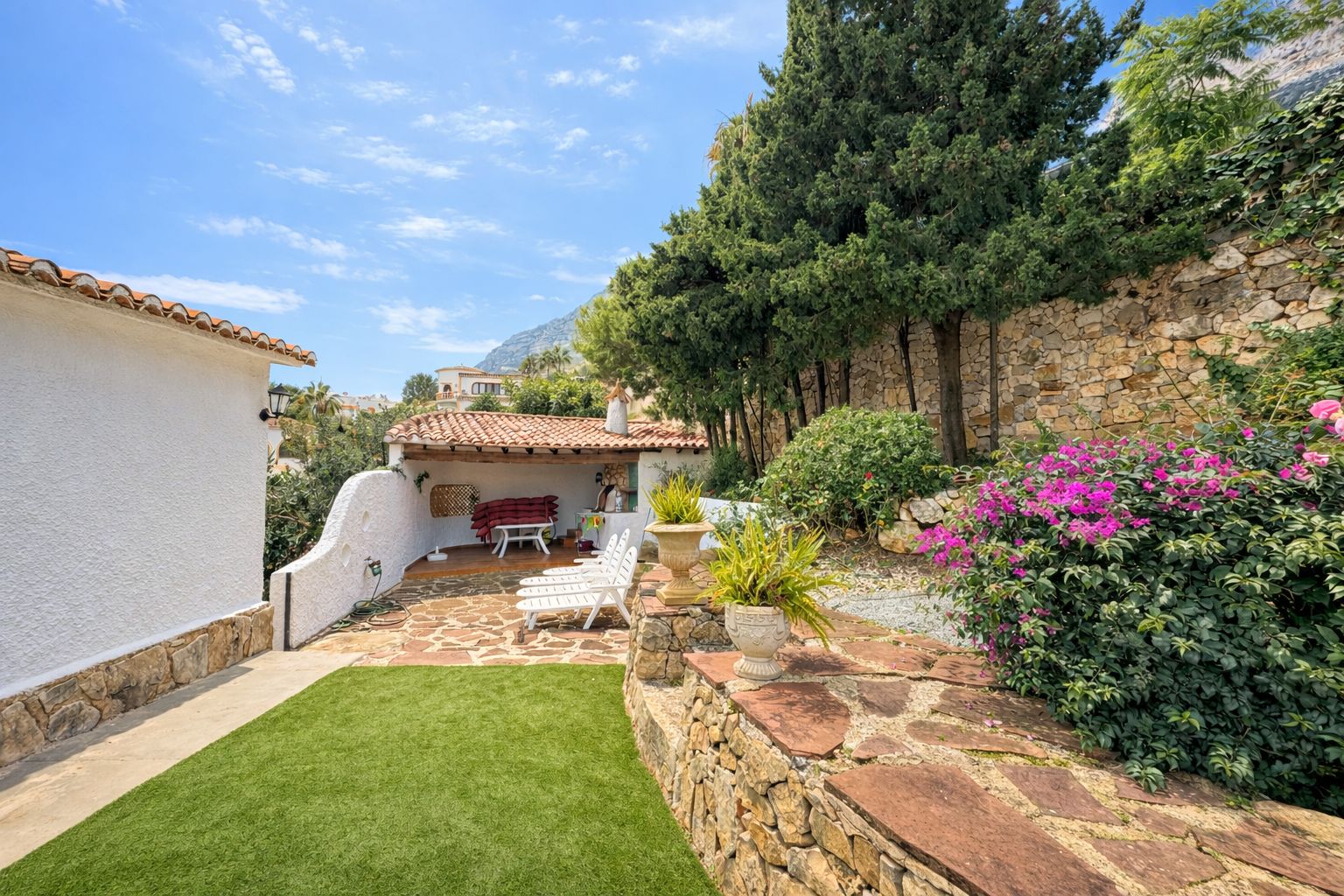Außenbereich mit Natursteinpflaster, Kunstrasen und überdachter Terrasse mit Ziegeldach. Bruchsteinmauern und mediterrane Bepflanzung säumen das Grundstück.