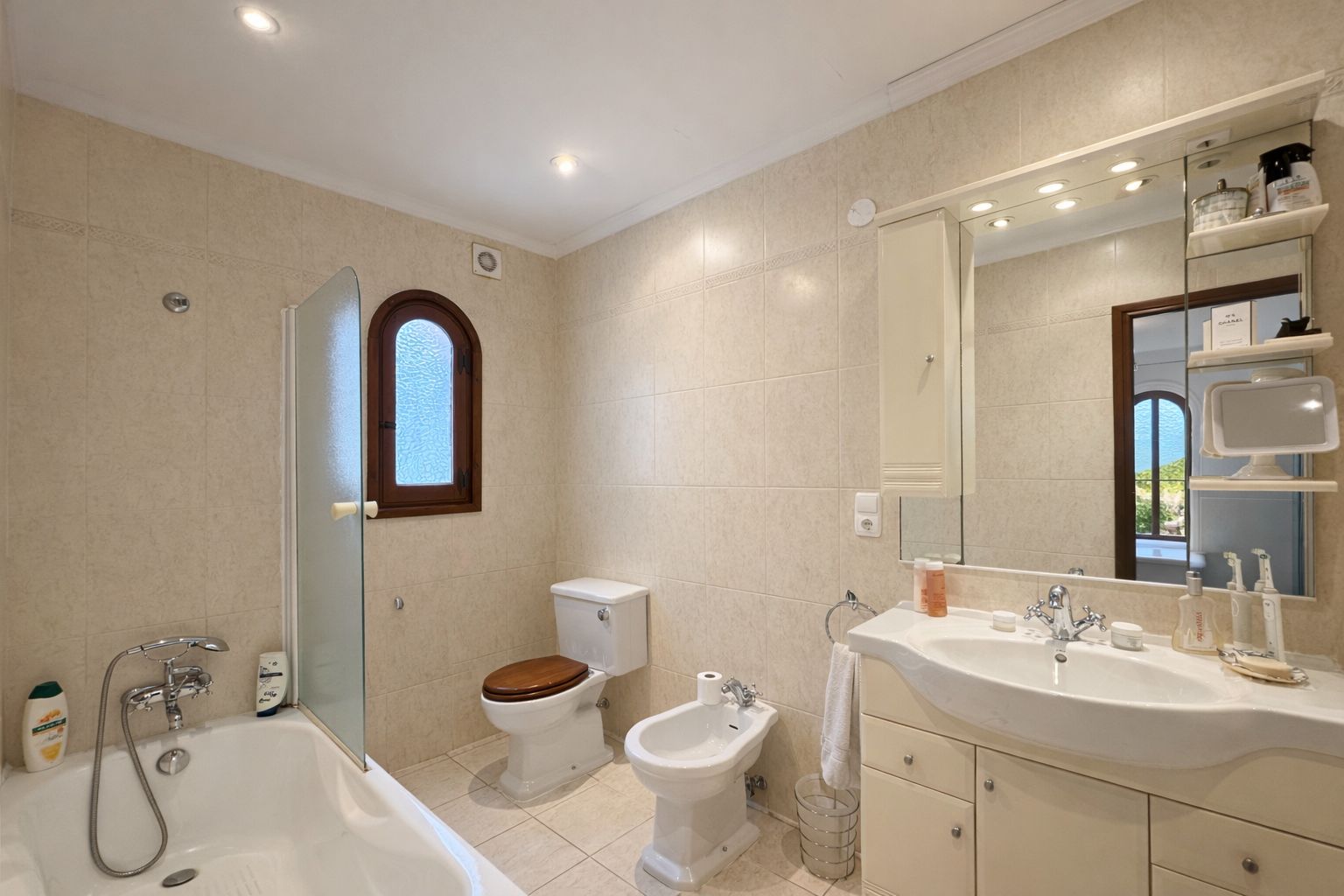 Badezimmer mit Badewanne, Glastrennwand, Bidet und WC mit Holzdeckel. Ausgestattet mit Waschtisch, großem Spiegel und einem Rundbogenfenster mit Holzrahmen.